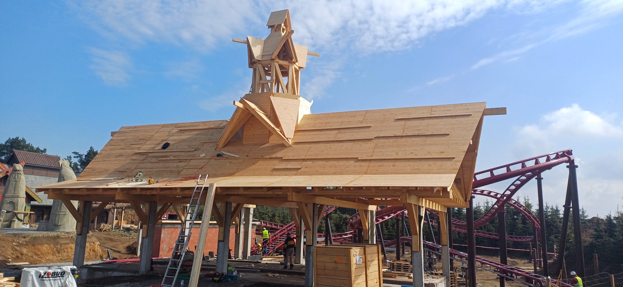 Konstrukcja dachu z drewna na etapie budowy, widoczna wieżyczka i elementy dekoracyjne. W tle fragment kolejki górskiej i błękitne niebo.