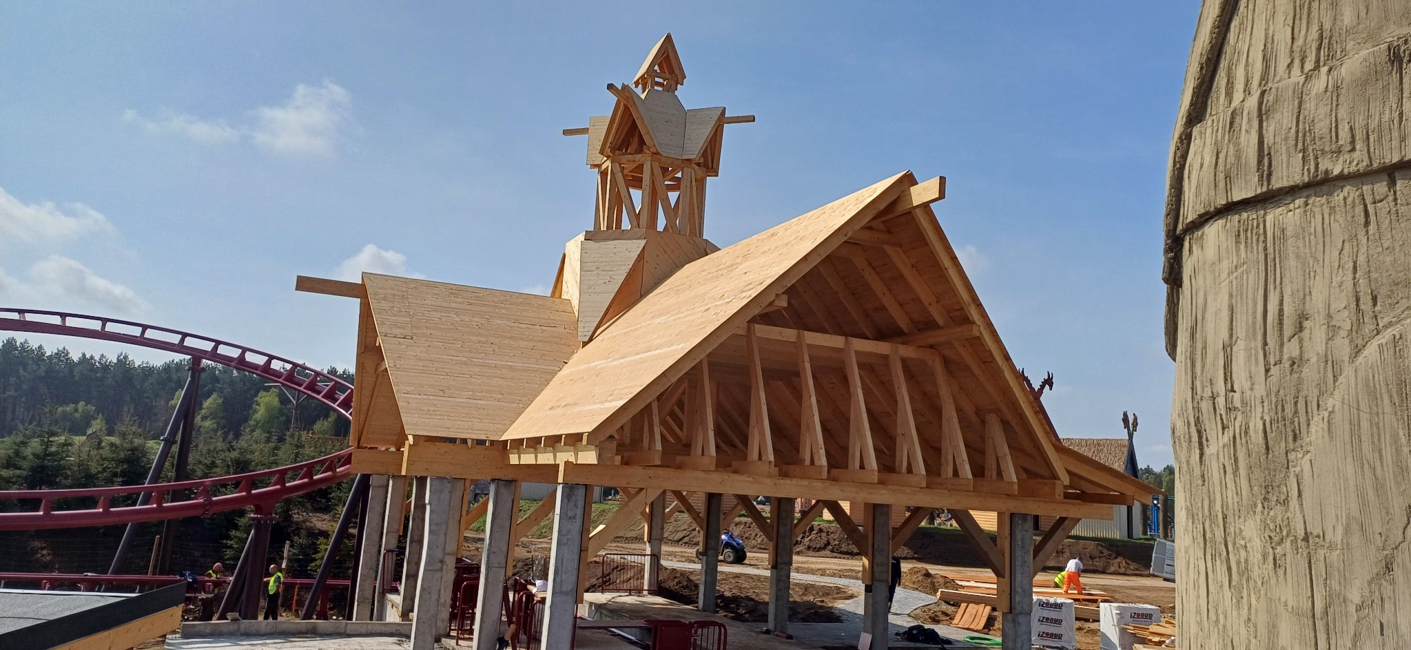 Wznoszenie drewnianej konstrukcji dachu z wieżą, na tle kolejki górskiej i dekoracyjnej skały. Widoczni pracownicy budowlani.