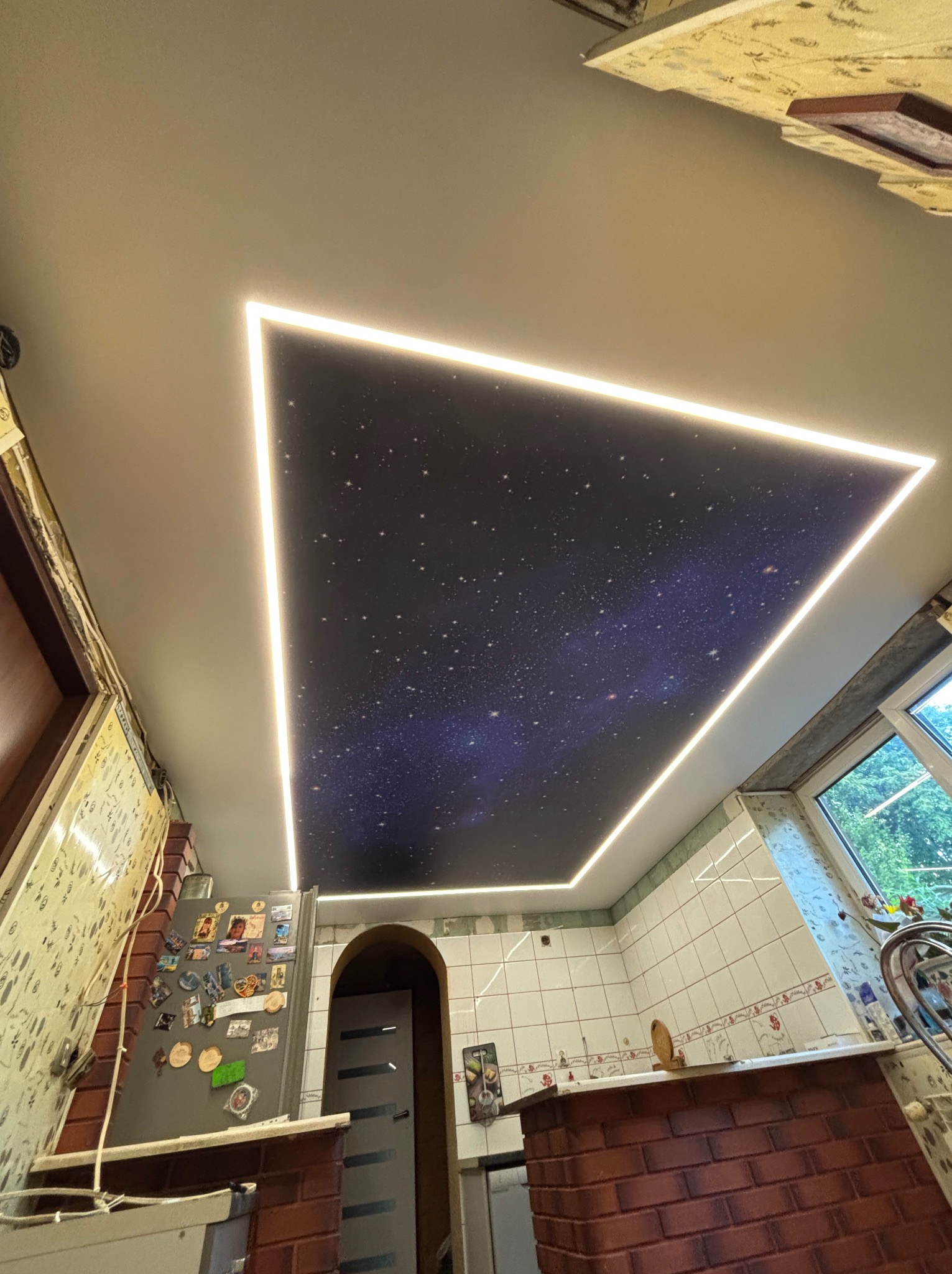 Nowoczesny sufit napinany w kuchni z motywem kosmosu, podświetlony ledowym obramowaniem w kształcie trójkąta. Widok z dołu.