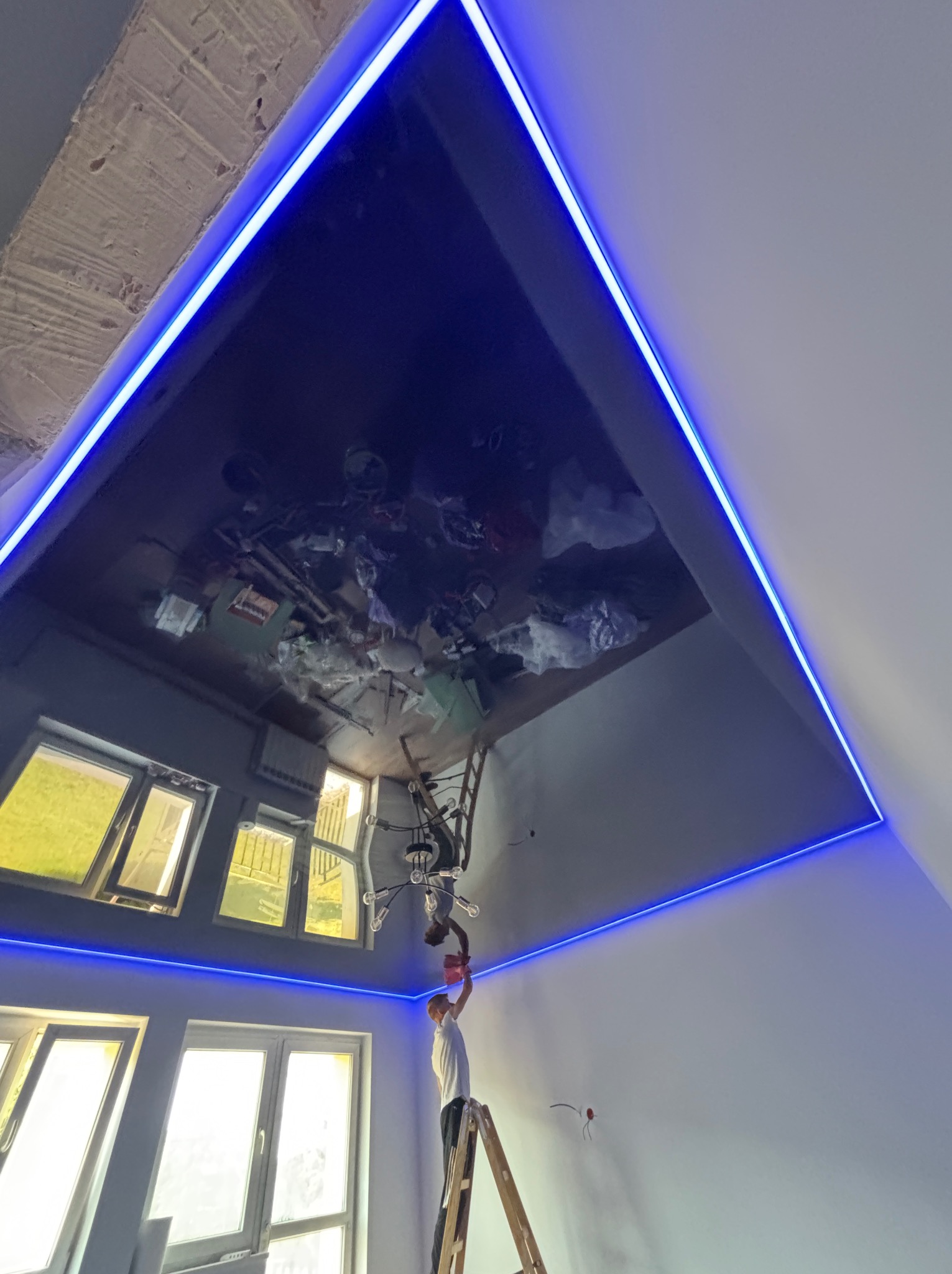 Błyszczący, ciemny sufit napinany z podświetleniem LED wzdłuż krawędzi, odbijający wnętrze pokoju z oknami i pracownikiem na drabinie podczas montażu.