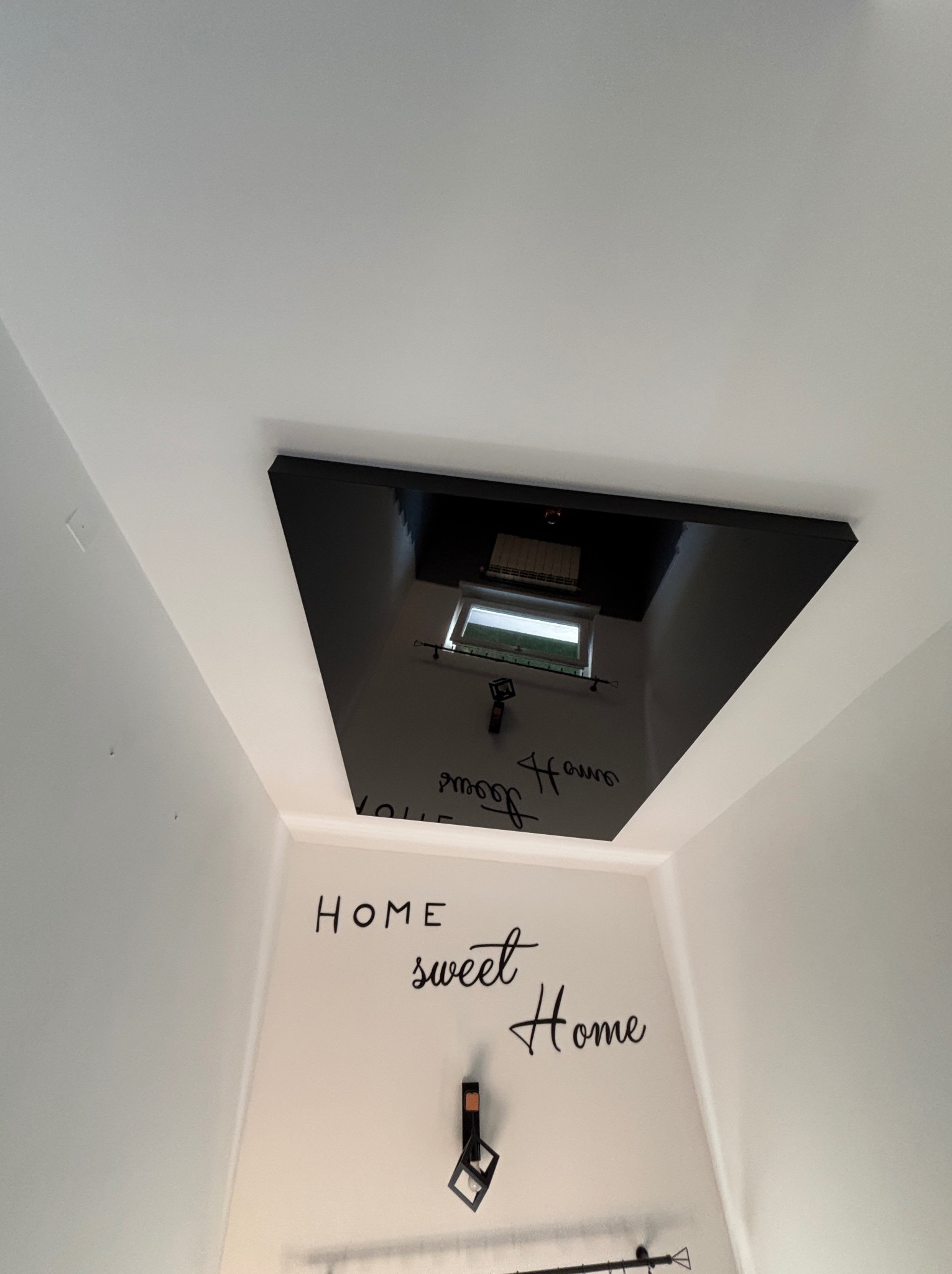Błyszczący, czarny sufit napinany z odbiciem okna i dekoracją ścienną 'Home sweet Home'. Ujęcie z dołu, podkreślające gładką powierzchnię sufitu.