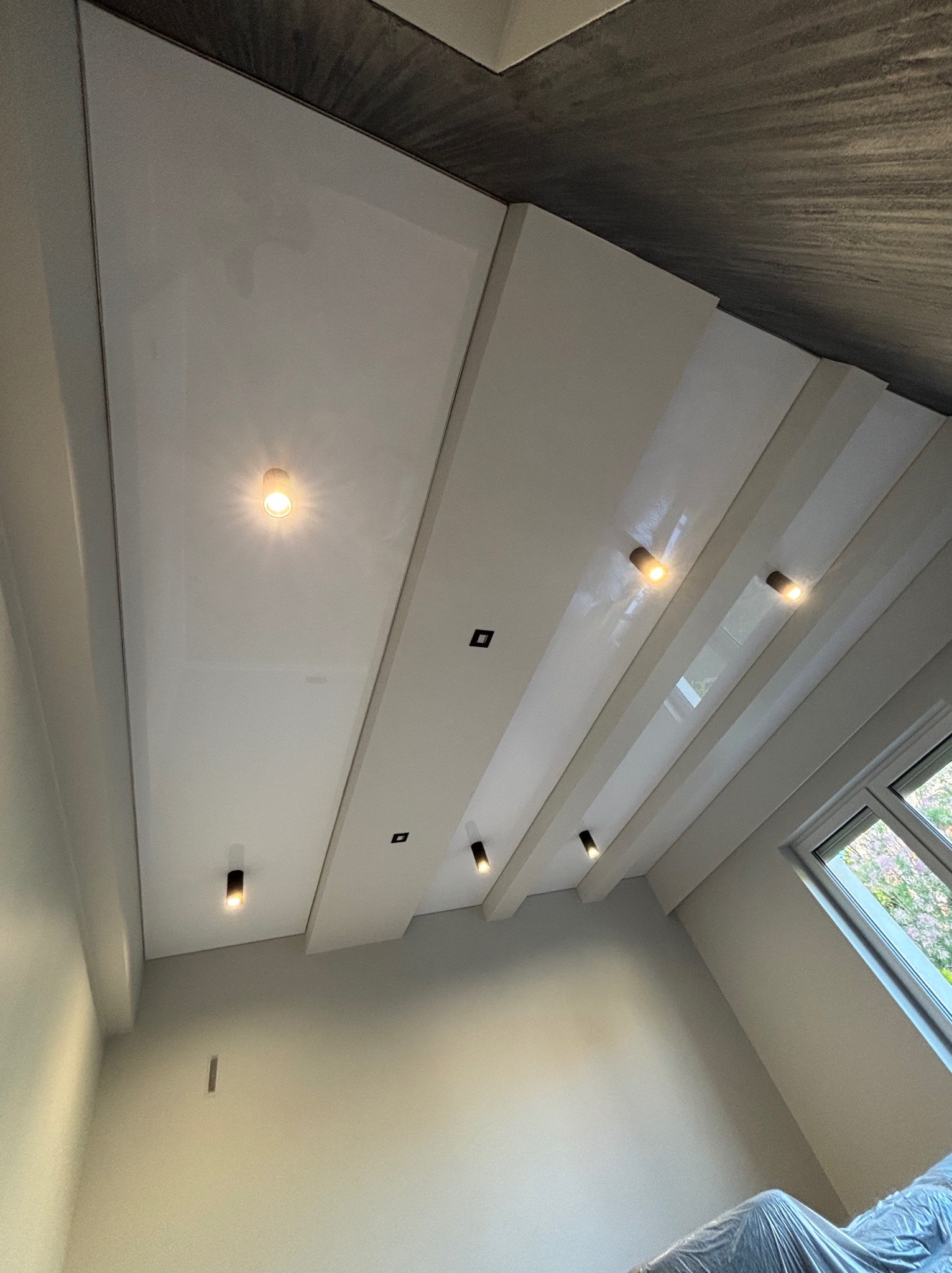 Biały, błyszczący sufit napinany z wbudowanym oświetleniem punktowym, widok z dołu pod kątem, fragment okna z widokiem na zewnątrz.