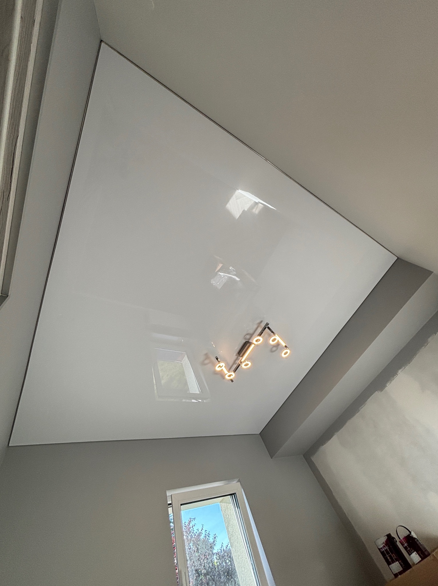 Błyszczący, biały sufit napinany z odbiciem lampy i okna. Widok z dołu, szare ściany, nowoczesne wykończenie wnętrza.