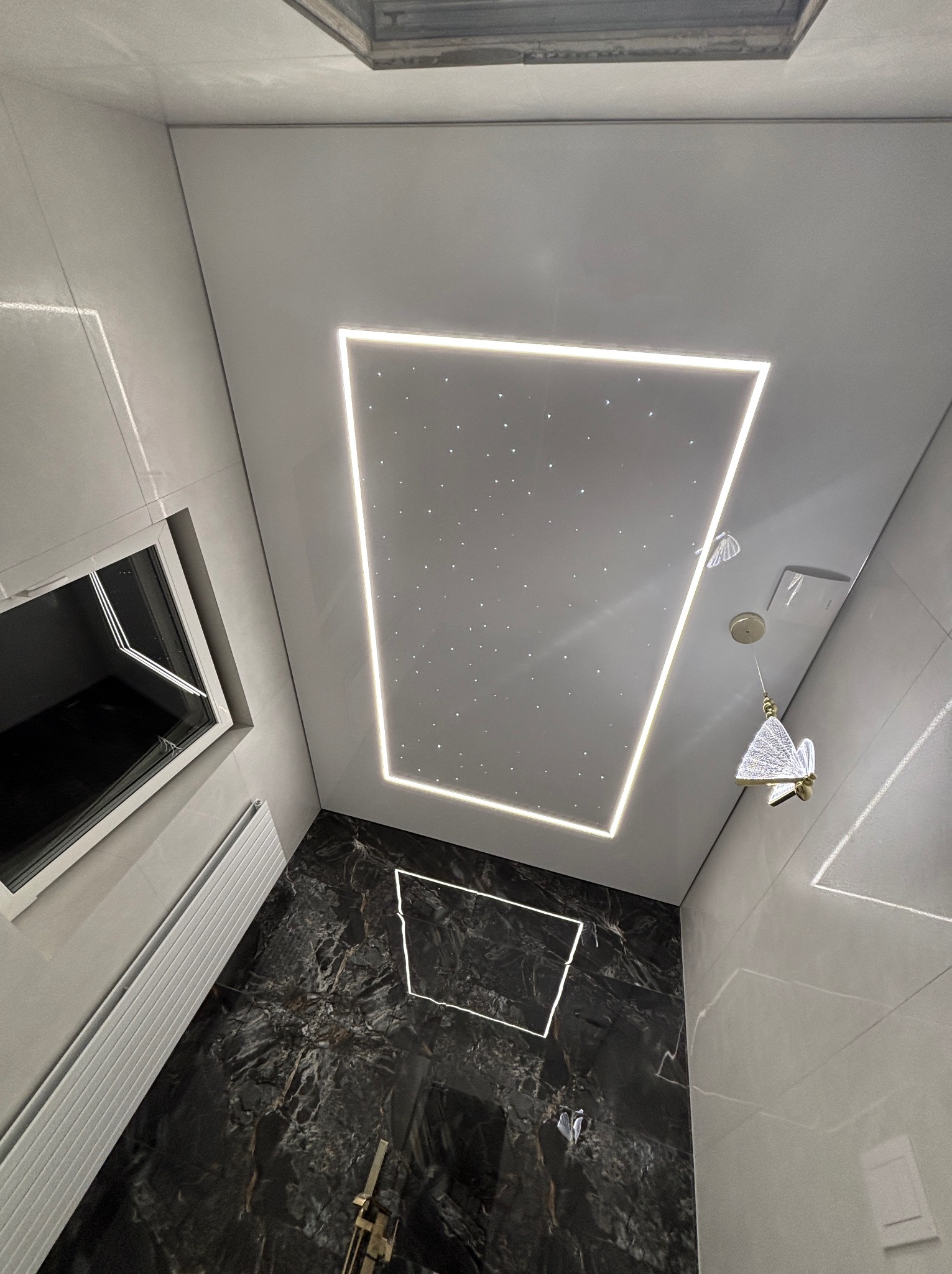 Nowoczesna łazienka z sufitem napinanym w kształcie prostokąta, podświetlonym diodami LED imitującymi gwiazdy. Czarno-białe marmurowe płytki na ścianach i podłodze.
