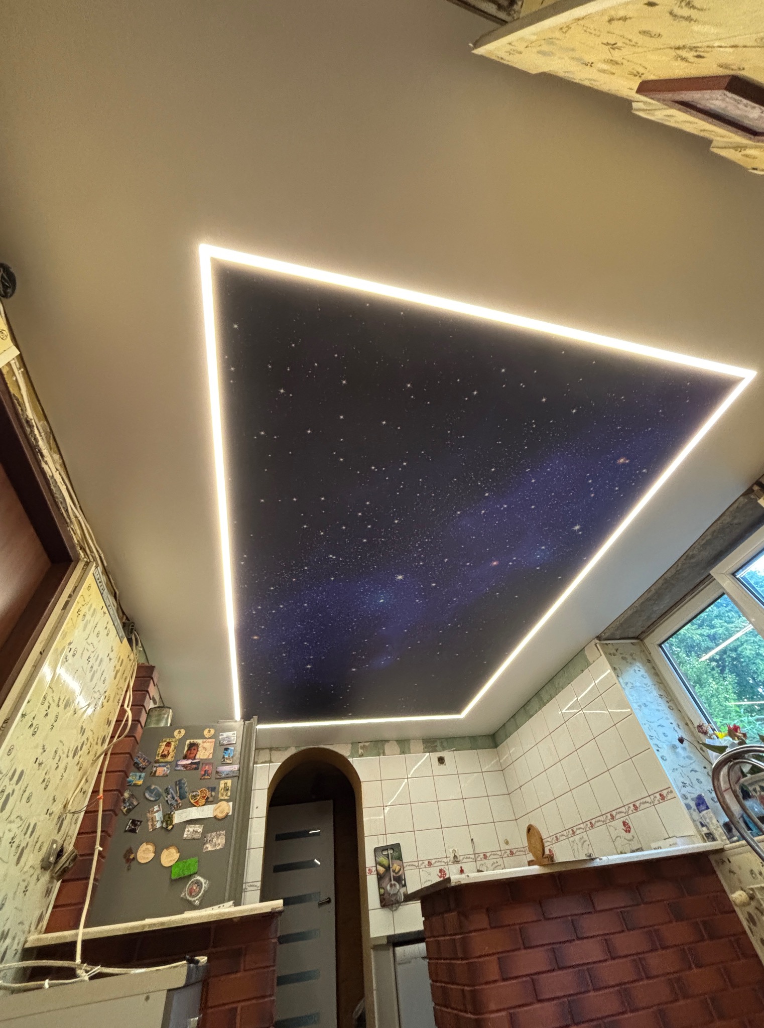 Sufit napinany z motywem kosmosu, podświetlony listwami LED w kształcie trójkąta, w kuchni z białymi płytkami i oknem z widokiem na zieleń.