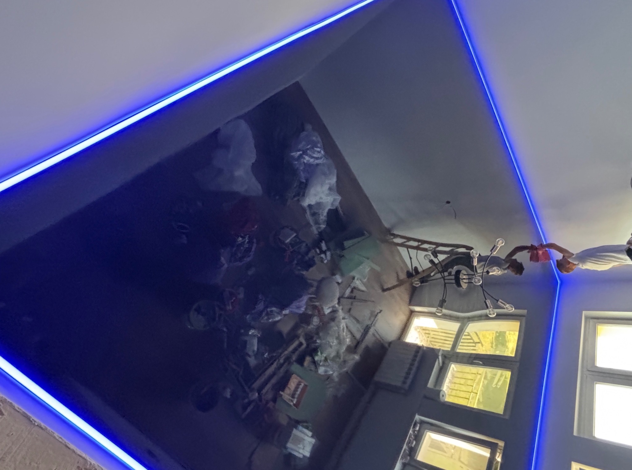 Błyszczący, granatowy sufit napinany z oświetleniem LED w Jastrząbiu. Widoczne odbicie wnętrza pokoju. Montażysta przy lampie.