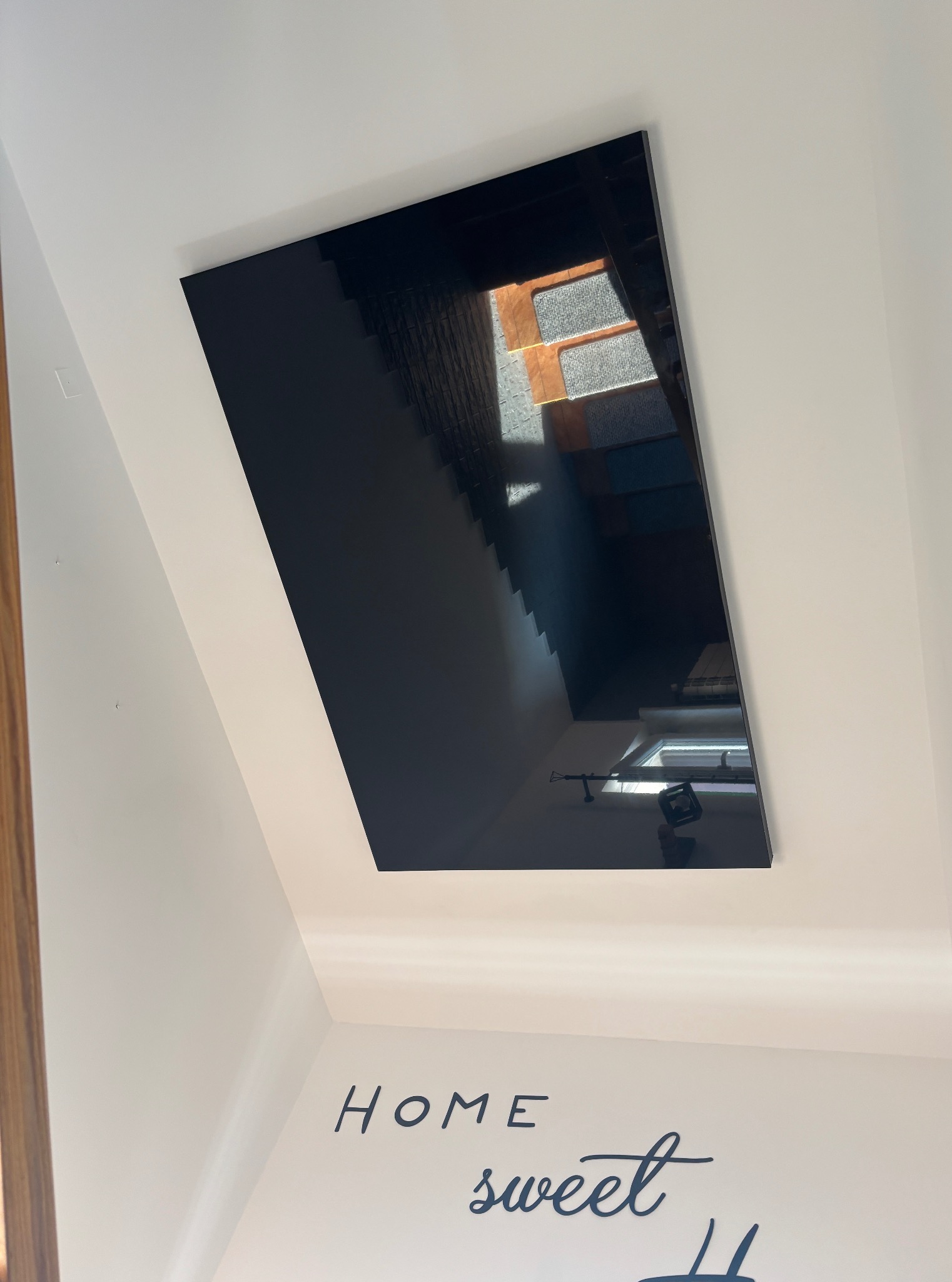 Błyszczący, czarny sufit napinany, odbijający schody i okno, zamontowany w białym pomieszczeniu z napisem 'Home sweet home'.