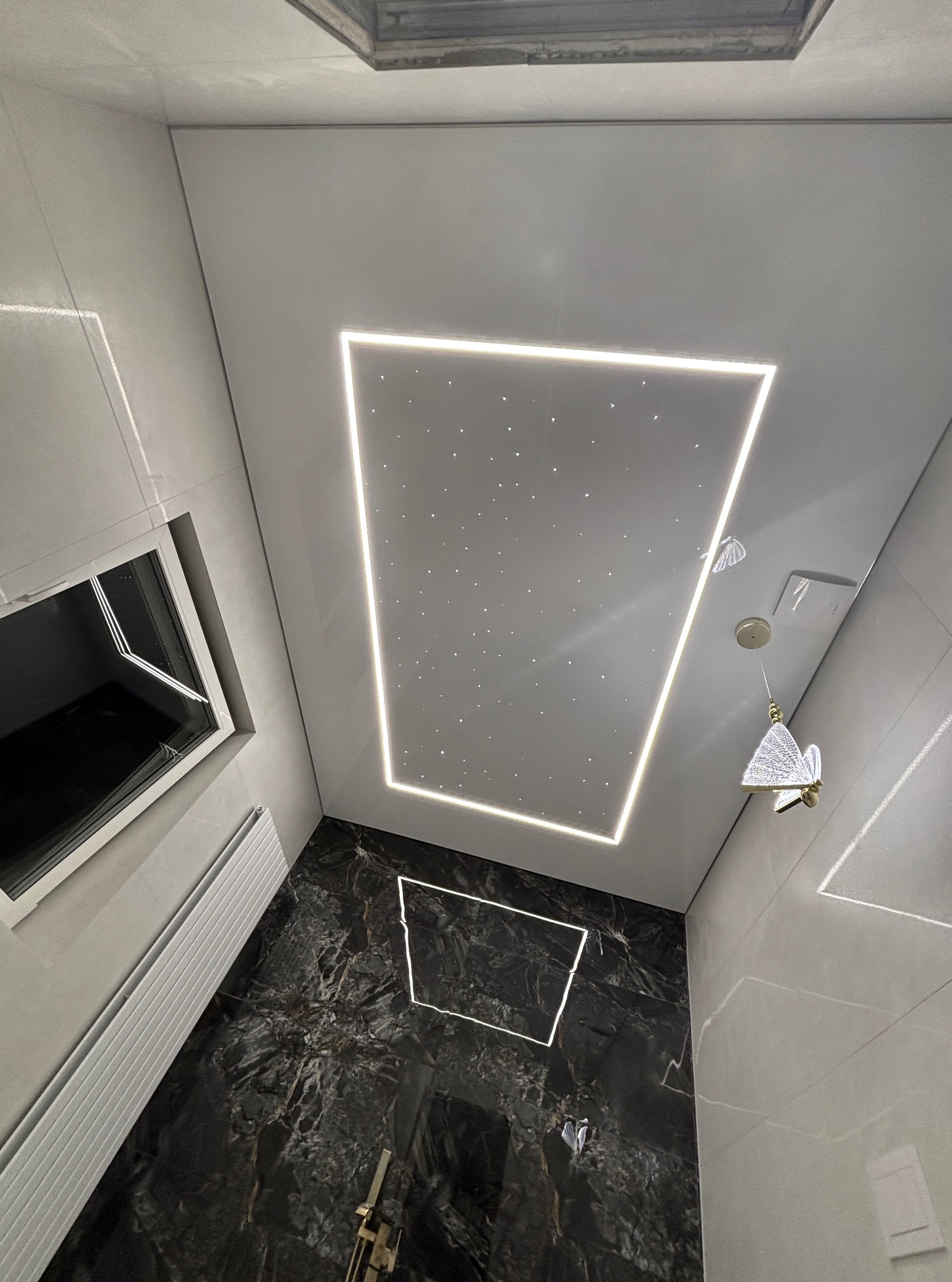 Nowoczesna łazienka z czarną marmurową podłogą i sufitem napinanym z oświetleniem LED imitującym gwiazdy, widziana z dołu, z lampą w kształcie motyla.