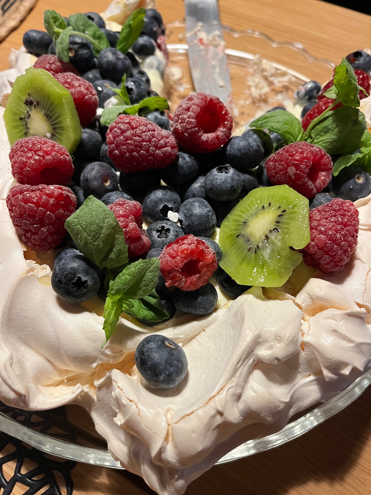 Pavlova z owocami: maliny, borówki, kiwi i mięta na bezie w szklanej misie. Część deseru została już zjedzona. Widoczny nóż. Idealny deser na catering.