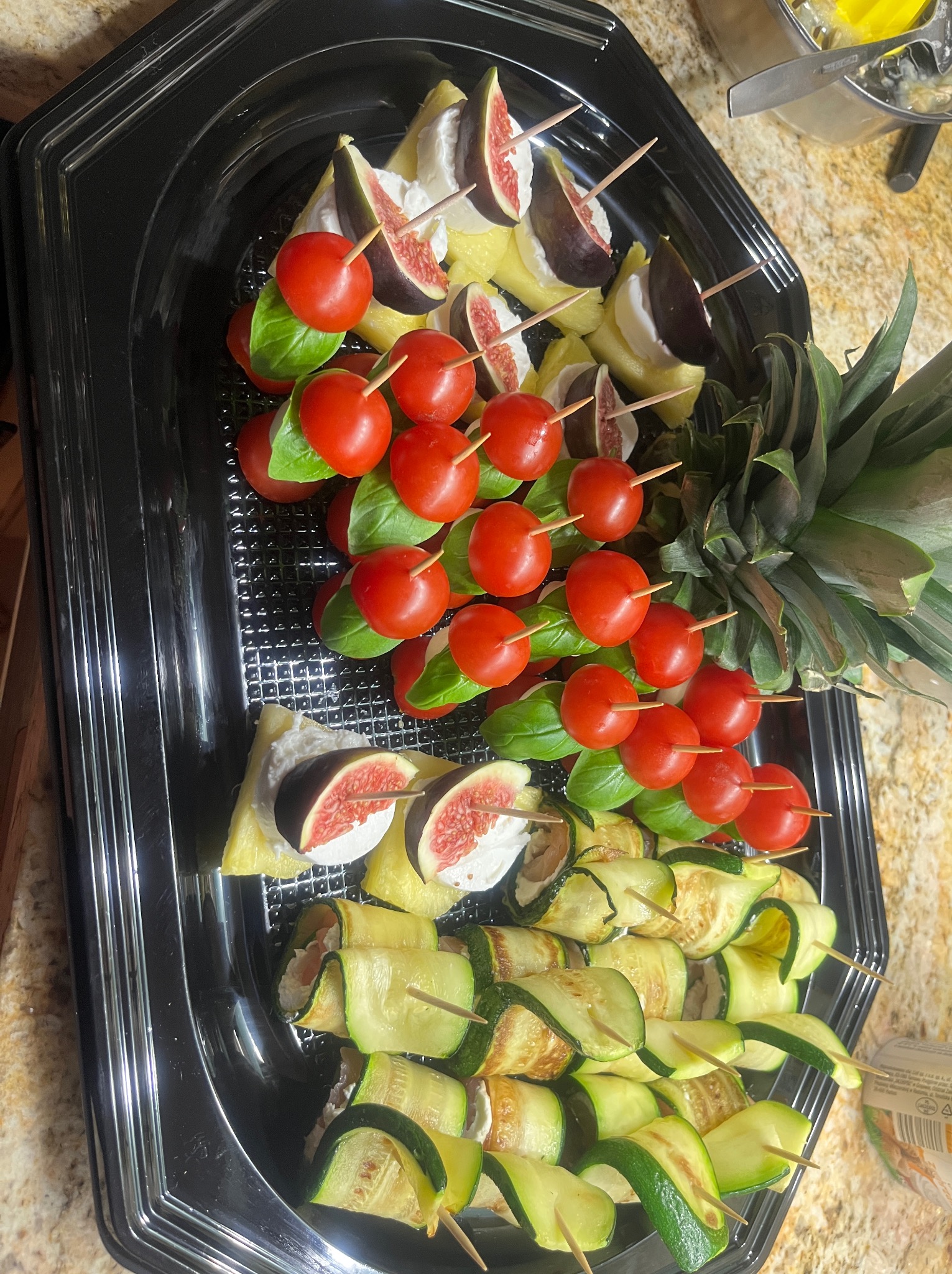 Elegancki catering: szaszłyki z fig, ananasa i sera, pomidorki cherry z bazylią i roladki z cukinii na czarnej tacy, obok ananas. Precyzyjne wykonanie, świeże składniki.