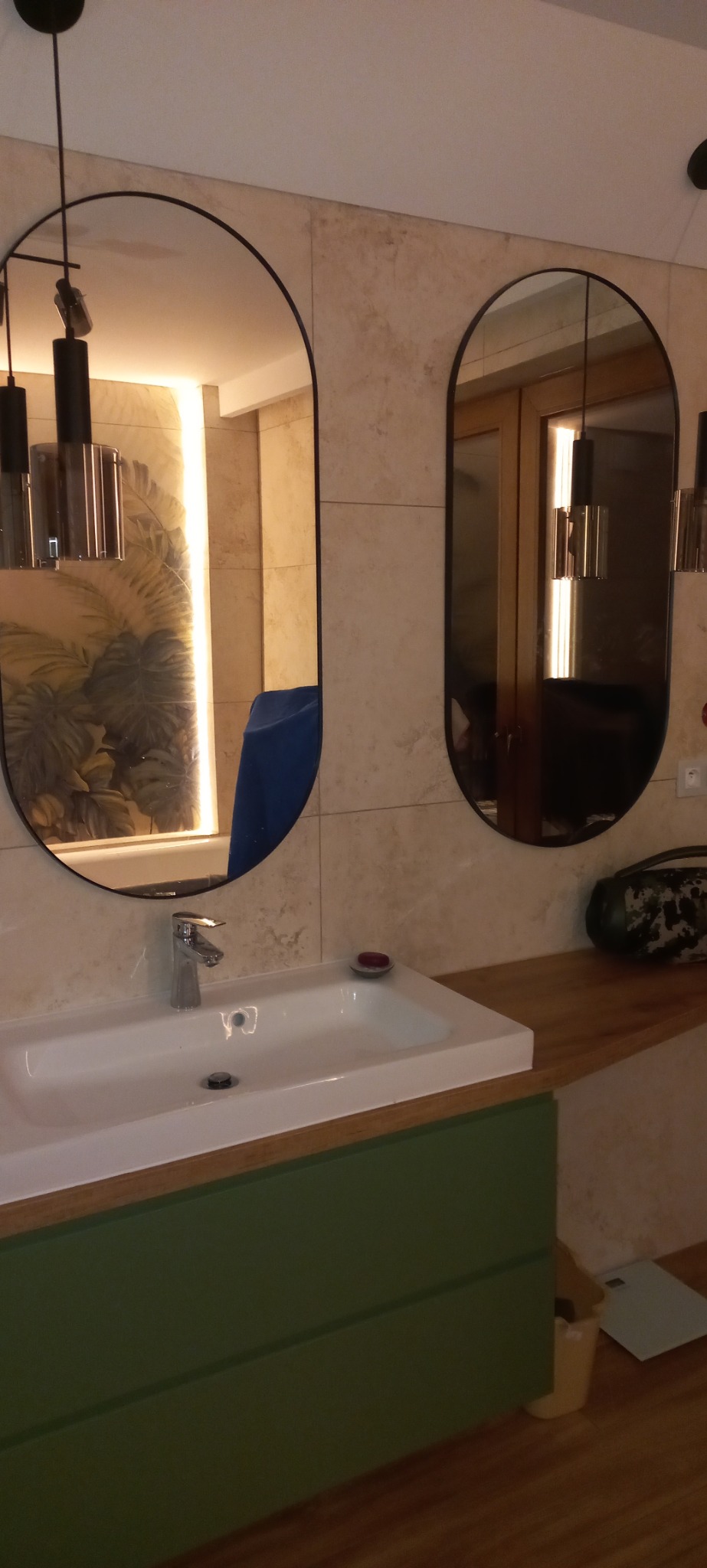 Nowoczesna łazienka z owalnymi lustrami, białą umywalką i zieloną szafką. Na ścianie beżowe płytki, a w lustrach odbijają się lampy i roślinny motyw.