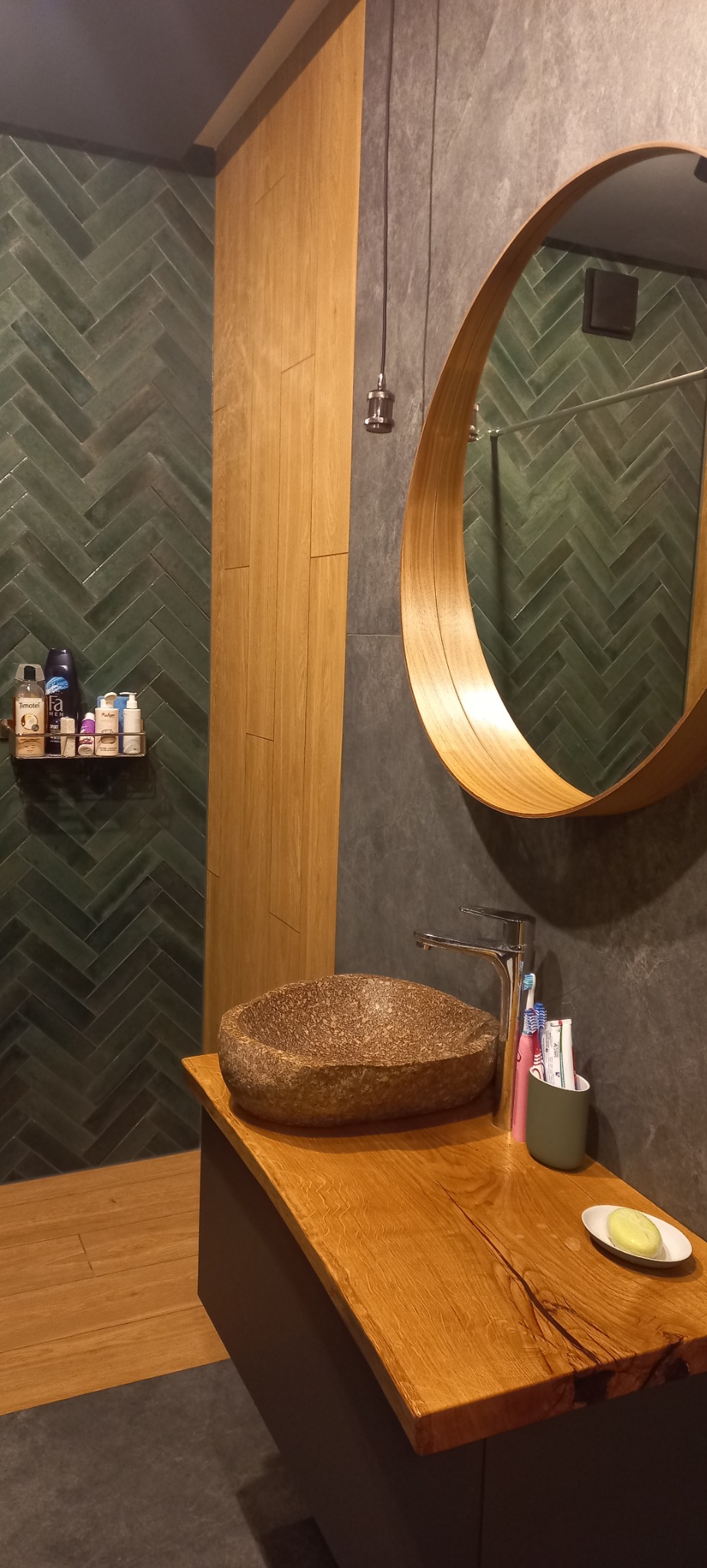 Nowoczesna łazienka z umywalką z kamienia na blacie z drewna, okrągłe lustro w drewnianej ramie, zielone płytki ułożone w jodełkę na ścianie.