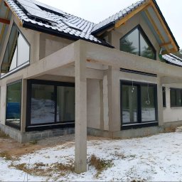 Urbański. Usługi budowlane. - Dom w stanie surowym zamkniętym, z oknami i dachem pokrytym czarną dachówką. Widoczne panele słoneczne na dachu i ślady śniegu na ziemi. Charakterystyczne trójkątne okno.