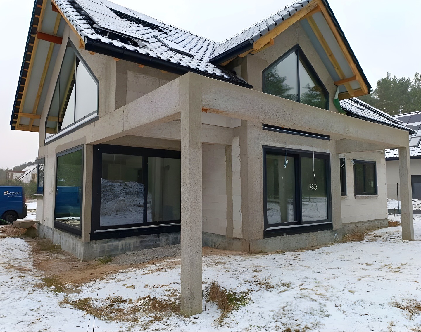 Dom w stanie surowym zamkniętym, z oknami i dachem pokrytym czarną dachówką. Widoczne panele słoneczne na dachu i ślady śniegu na ziemi. Charakterystyczne trójkątne okno.