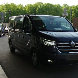 BusVoyage24 - Czarny Renault Trafic z włączonymi światłami, zaparkowany na ulicy w słoneczny dzień. Widoczny kierowca wewnątrz pojazdu i inne auta w tle.