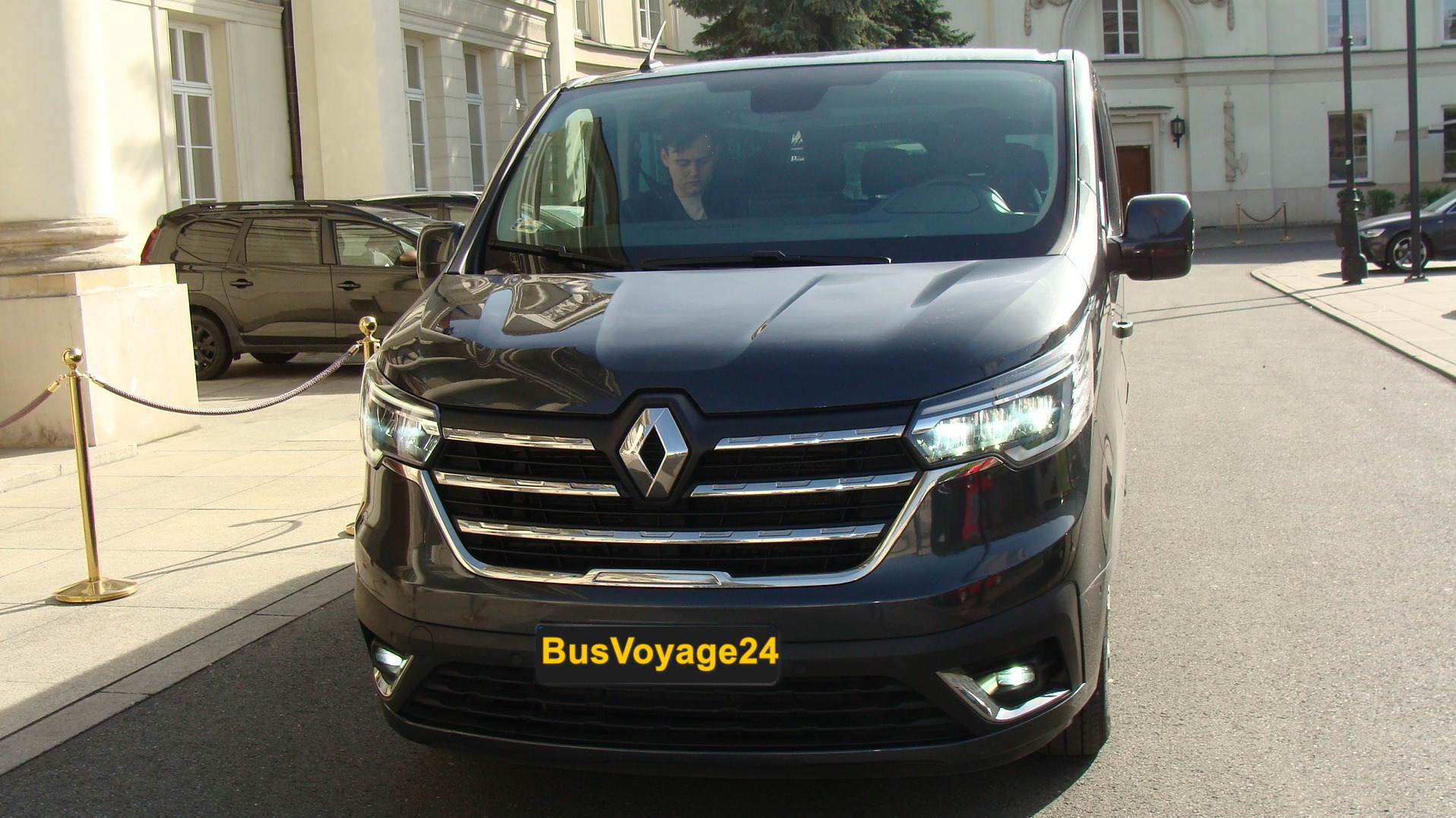 Czarny bus Renault Trafic z kierowcą za szybą, zaparkowany na ulicy przed budynkiem. Widoczny numer rejestracyjny BusVoyage24.