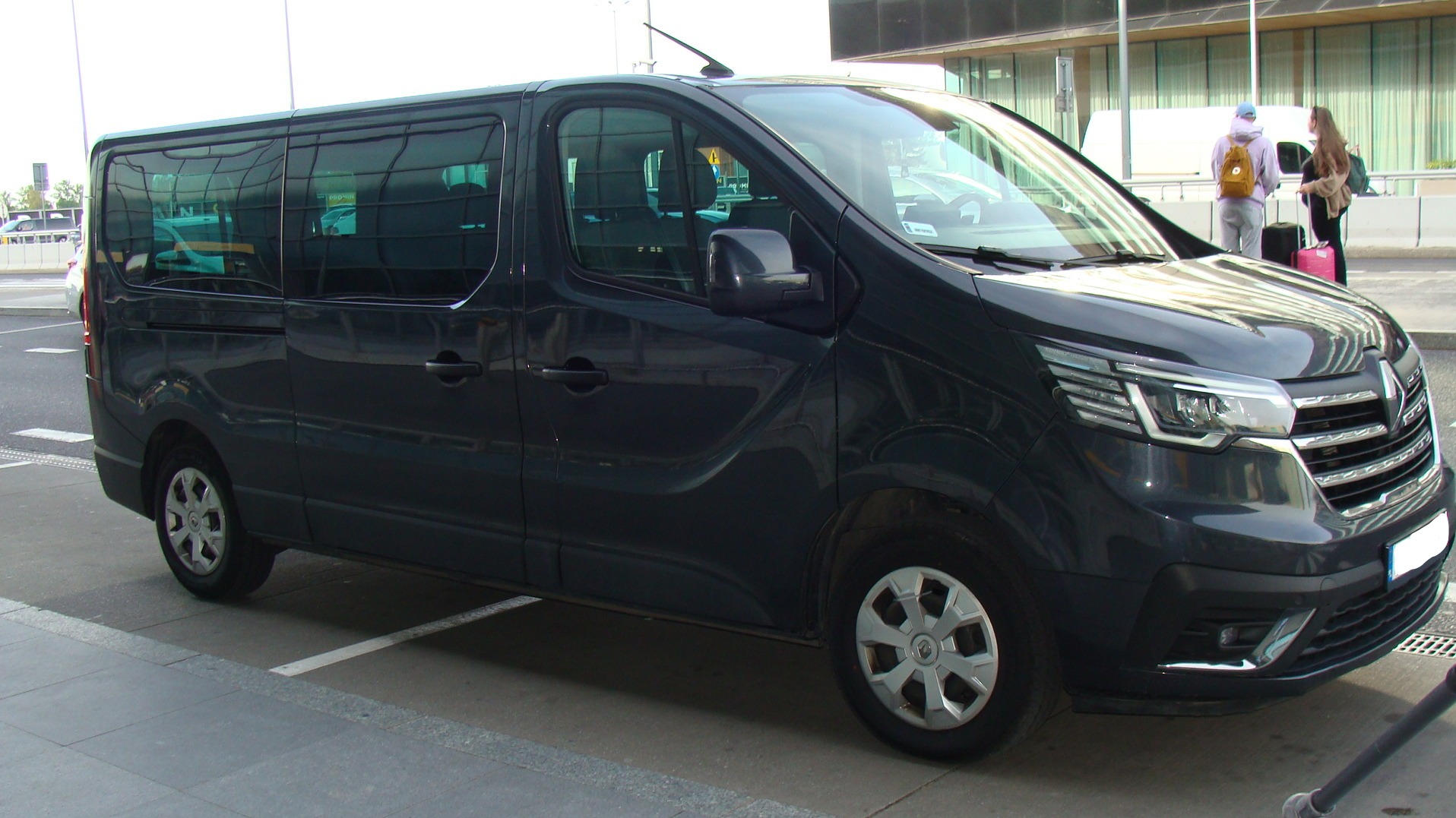 Ciemny bus Renault Trafic z pasażerami i bagażami w tle, zaparkowany na parkingu. Widok z boku, lekko od przodu. Światło dzienne.