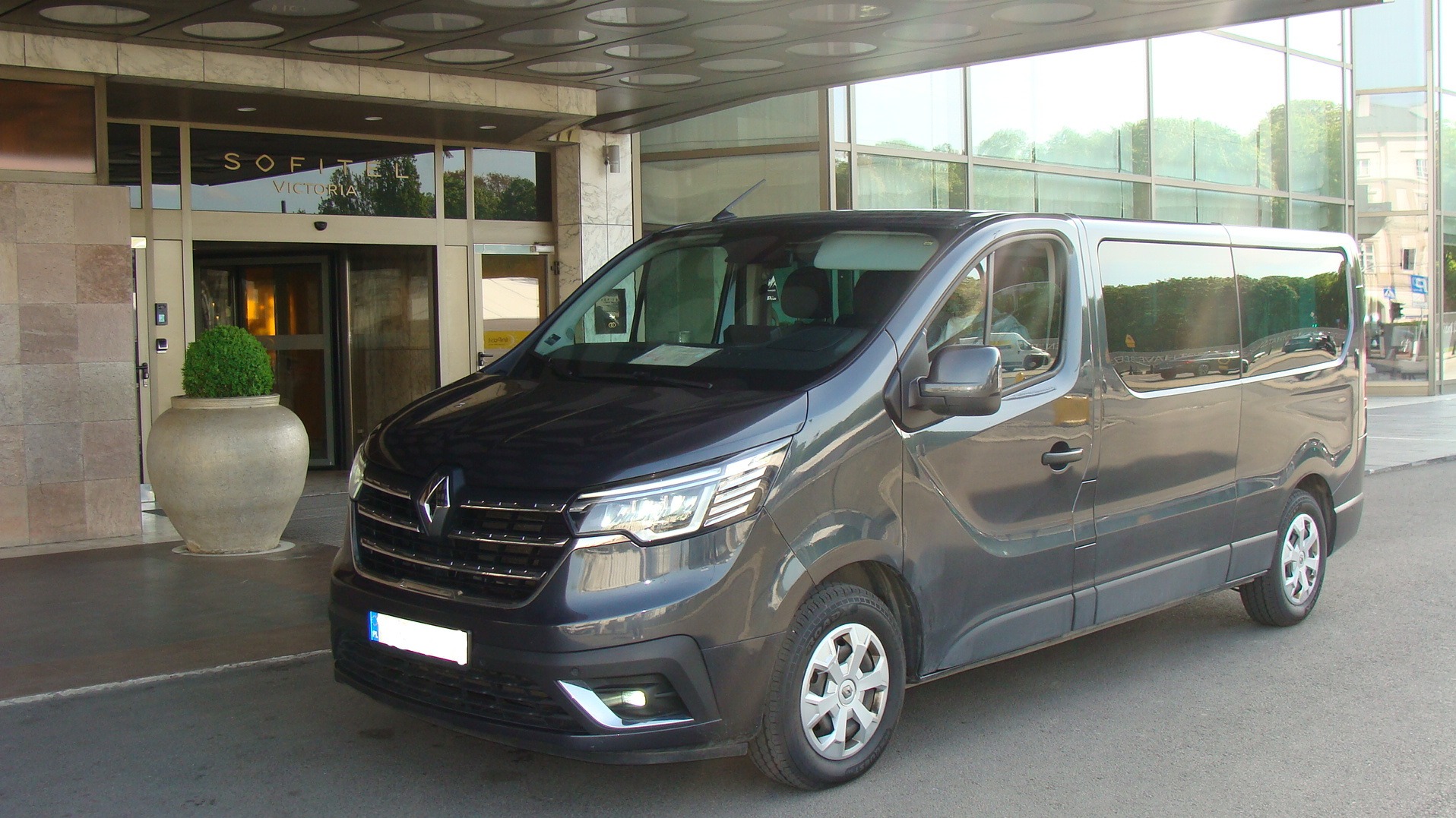 Szary bus Renault zaparkowany przed wejściem do hotelu Sofitel Victoria, z widocznym odbiciem otoczenia w szybach. Auto gotowe do transportu osób.