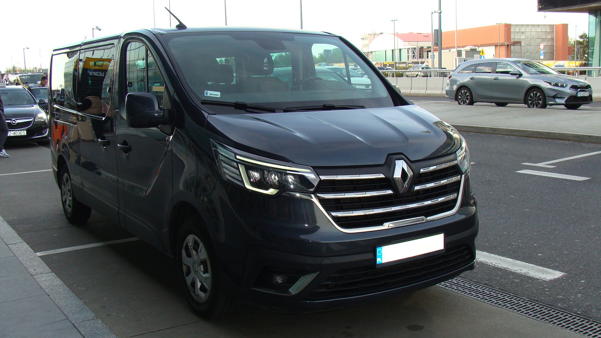 Ciemny bus Renault Trafic z przodu, zaparkowany na parkingu z innymi samochodami w tle. Widoczne detale karoserii i reflektory. Tablica rejestracyjna zasłonięta.