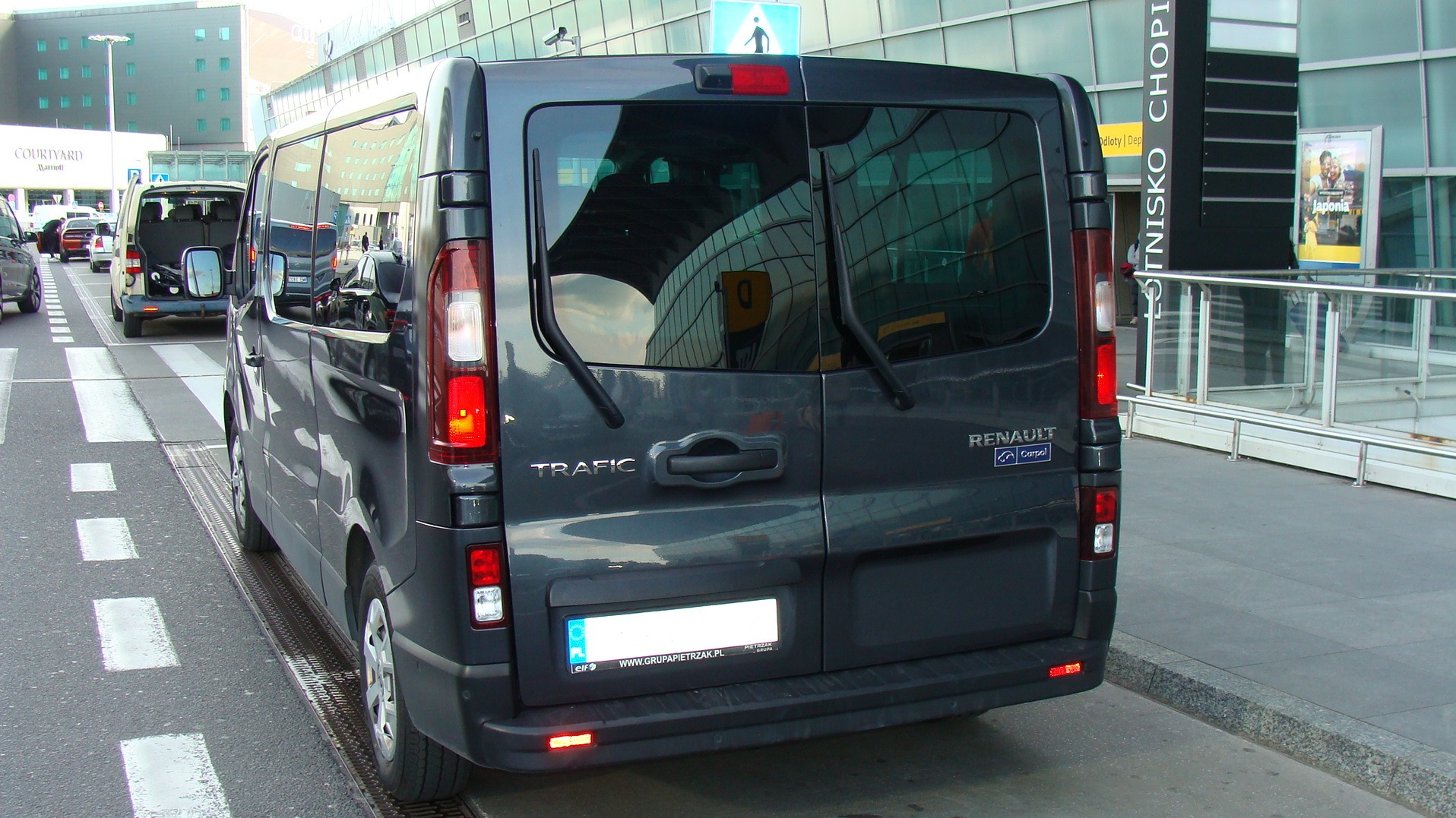 Szary bus Renault Trafic z tyłu, zaparkowany na pasach dla pieszych przed lotniskiem Chopina w Warszawie. Widoczne budynki lotniska i inne pojazdy.