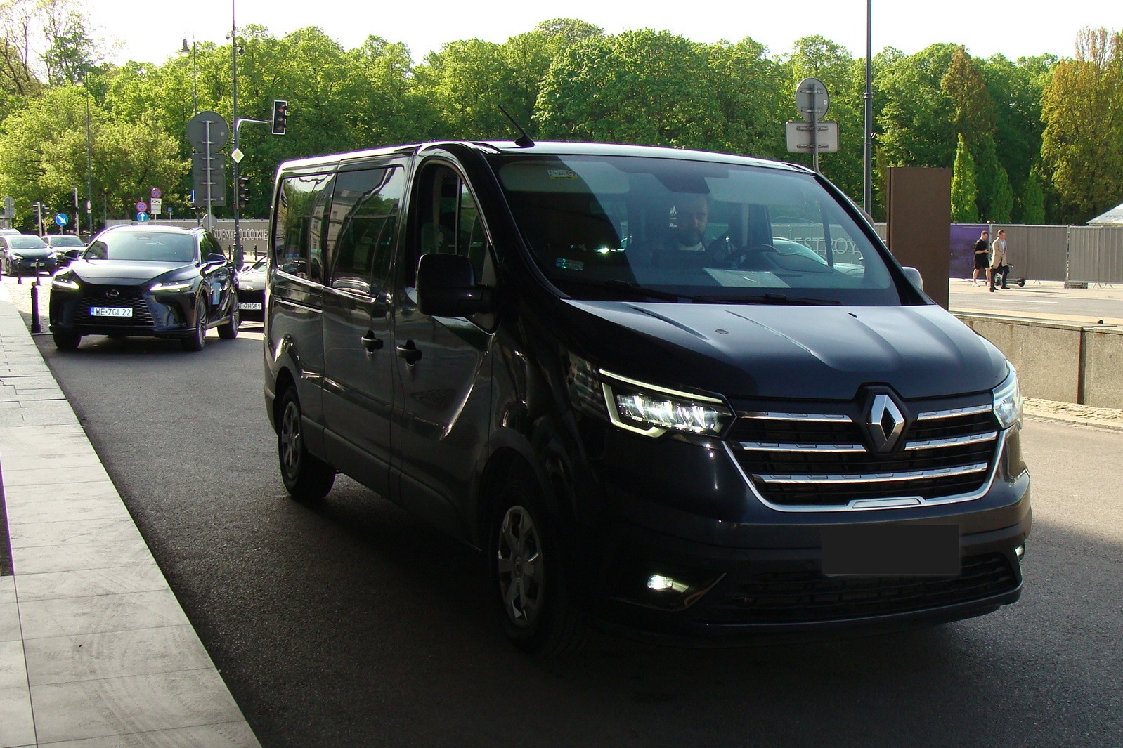 Czarny Renault Trafic z włączonymi światłami, zaparkowany na ulicy w słoneczny dzień. Widoczny kierowca wewnątrz pojazdu i inne auta w tle.