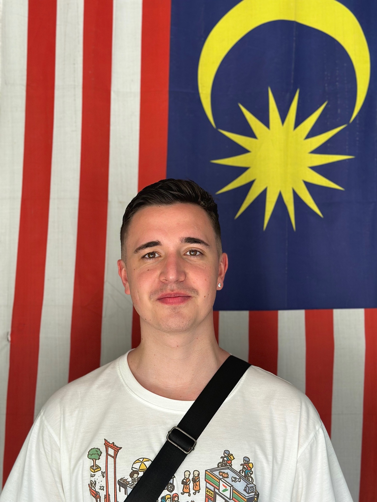 Portret mężczyzny na tle flagi Malezji. Ubrany w białą koszulkę z graficznym wzorem i czarną torbę na ramię. Uśmiechnięty, z kolczykiem w lewym uchu.
