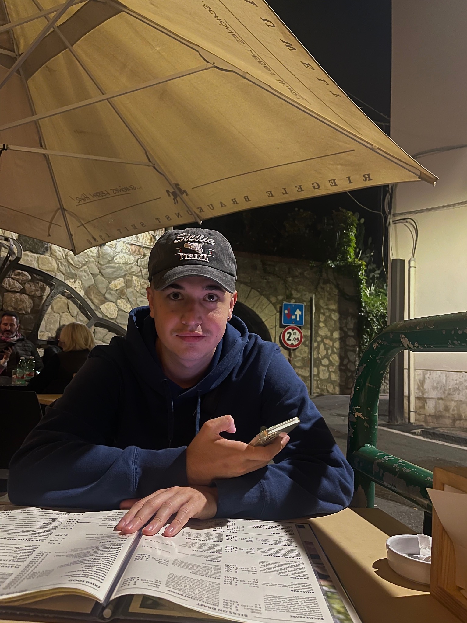 Młody mężczyzna w czapce 'Sicilia' siedzi przy stoliku w restauracji pod parasolem z menu przed sobą i telefonem w dłoni. W tle kamienna ściana.