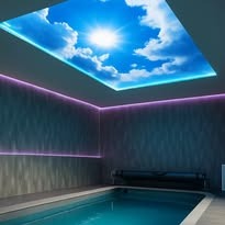 Błękitne niebo z promieniami słońca na suficie napinanym nad krytym basenem, oświetlenie LED wzdłuż ścian, nowoczesne wykończenie.
