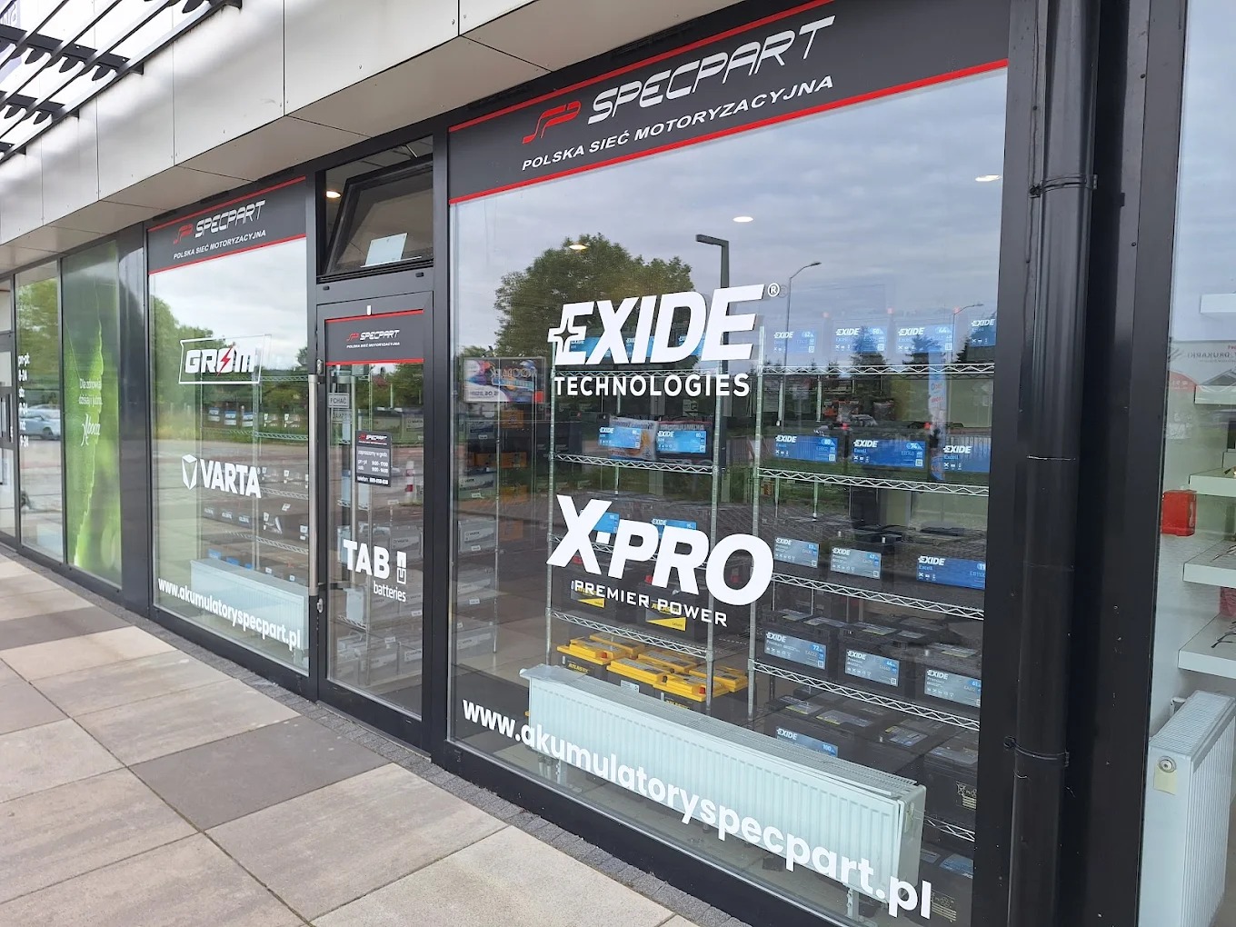 Fasada sklepu motoryzacyjnego Specpart z logo Exide i Varta na szybach, prezentująca szeroki wybór akumulatorów na półkach wewnątrz.