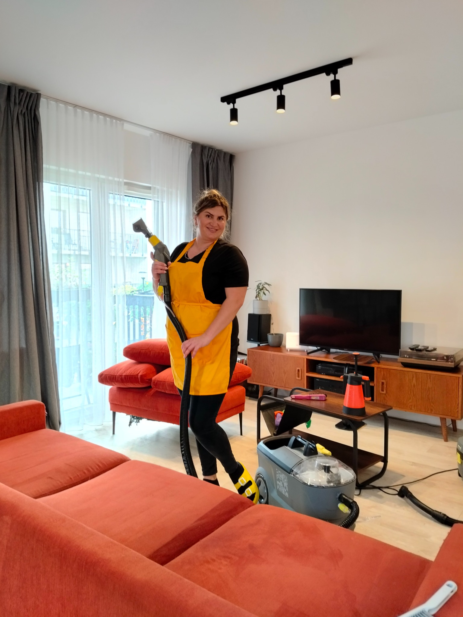 Uśmiechnięta kobieta w żółtym fartuchu prezentuje odkurzacz piorący Karcher w salonie z pomarańczową sofą i telewizorem. Usługa czyszczenia tapicerki.