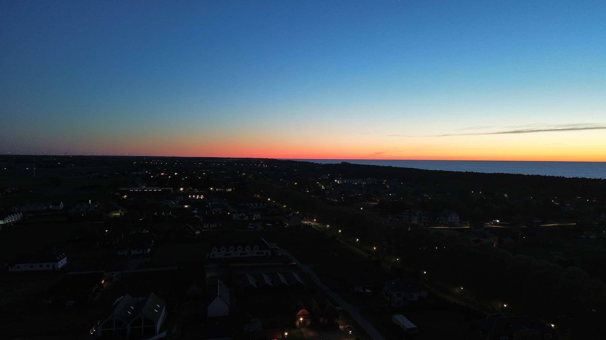 Nocny widok z lotu ptaka na miasto nad morzem, oświetlone latarniami, z pomarańczowo-niebieskim niebem o zmierzchu. Widoczne budynki, drzewa i linia horyzontu.