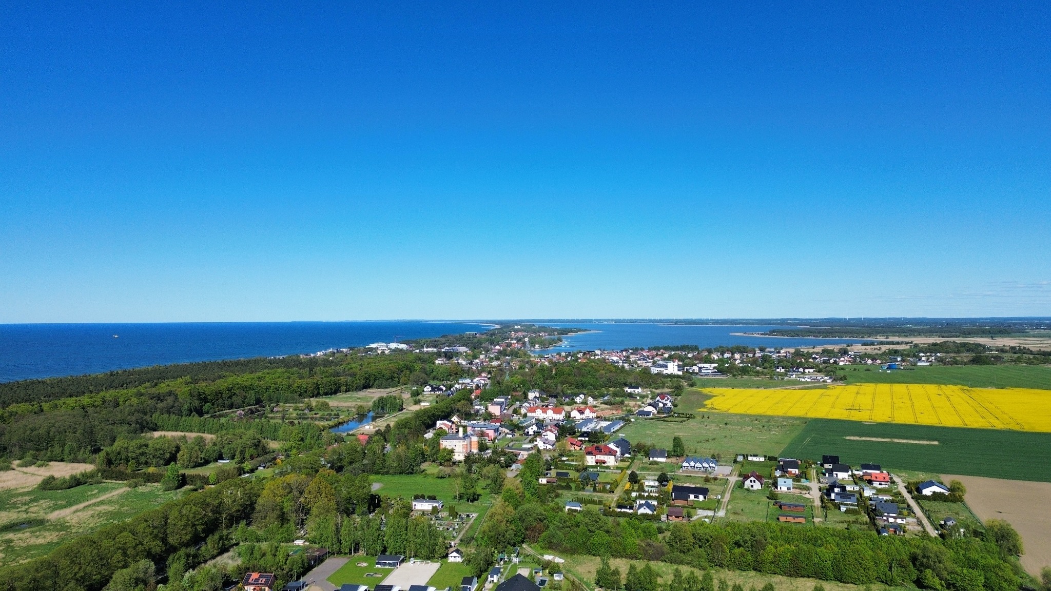 Panorama z lotu ptaka: błękit morza i nieba nad zielenią lasów, pól i zabudowań małego miasteczka. W tle widoczne turbiny wiatrowe.