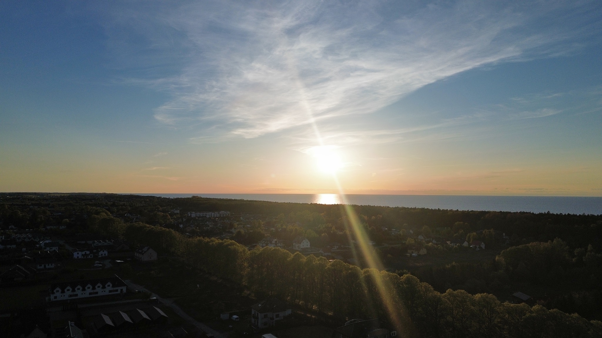 Pejzaż z lotu ptaka: zachód słońca nad morzem, w dole widoczne domy i drzewa. Promienie słońca odbijają się w wodzie, tworząc złoty blask.