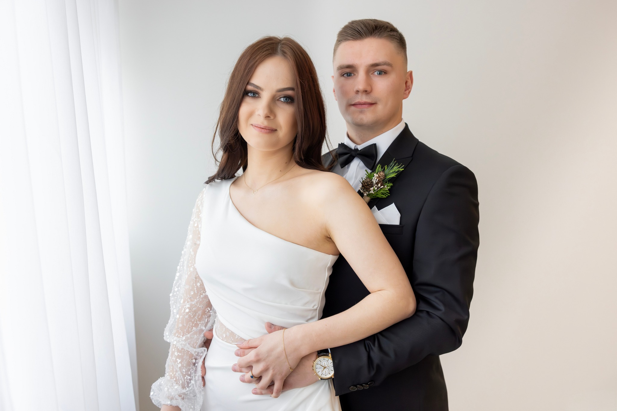 Elegancka para młoda: brunetka w asymetrycznej sukni z koronkowym rękawem, obejmowana przez mężczyznę w czarnym smokingu z butonierką. Jasne tło, naturalne światło.