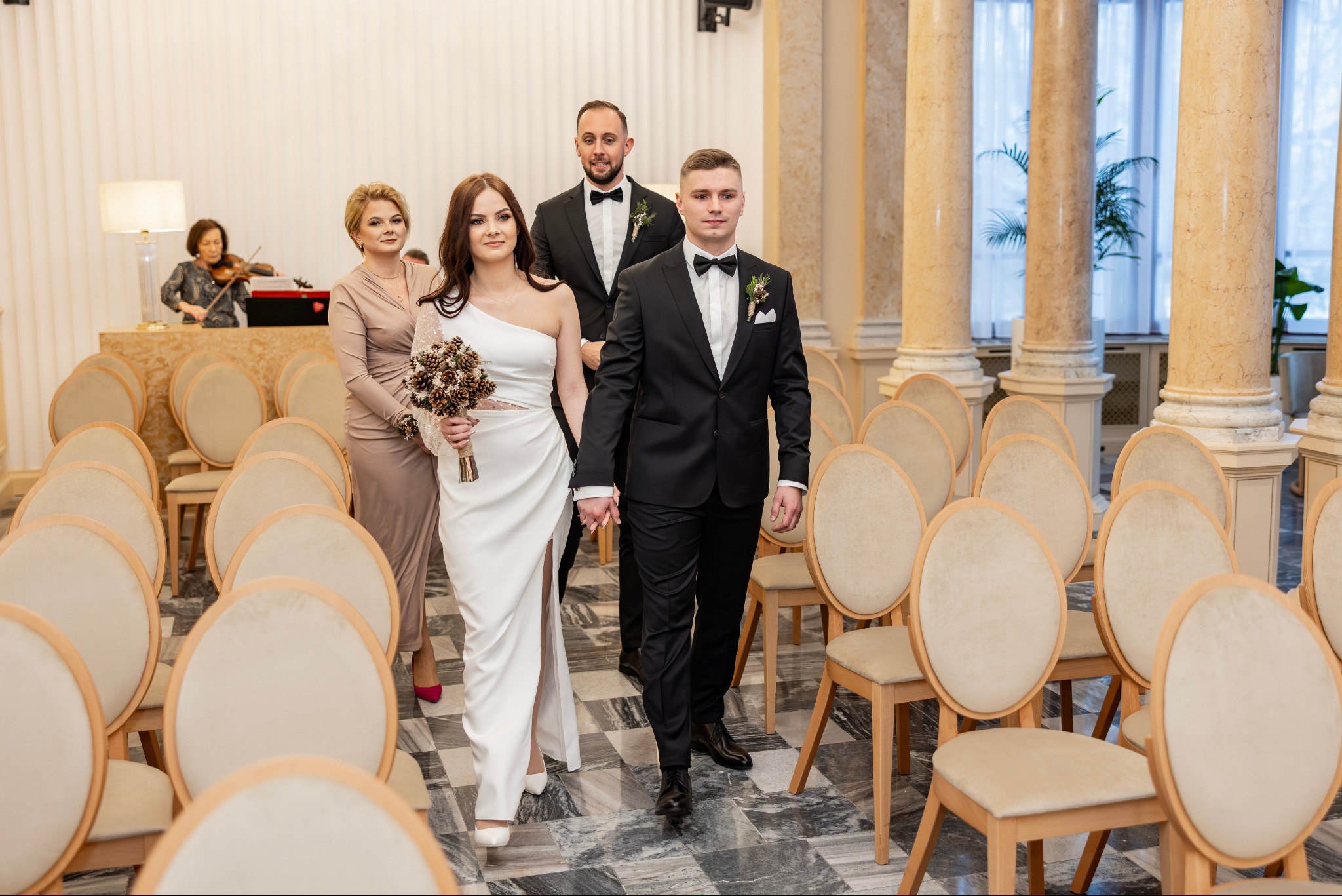 Para młoda w eleganckich strojach wchodzi do sali ślubnej, trzymając się za ręce. W tle goście i skrzypaczka. Sala z kolumnami i krzesłami dla gości.