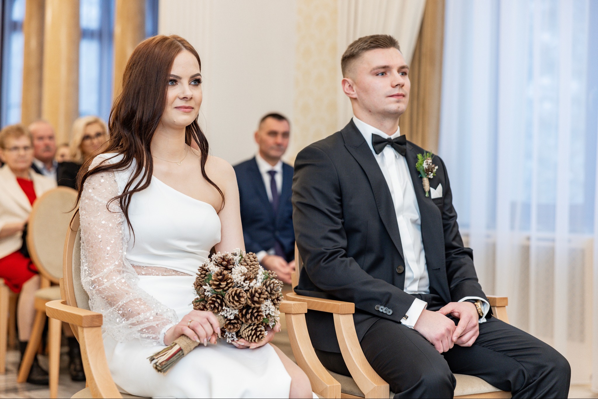 Para młoda siedzi podczas ceremonii ślubnej. Panna młoda w białej sukni z bukietem z szyszek, pan młody w czarnym garniturze z muchą. W tle goście.
