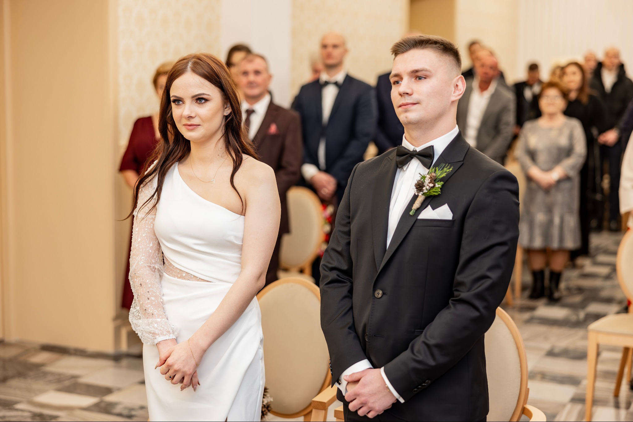 Para młoda, panna młoda w białej sukni z jednym rękawem z cekinami, pan młody w czarnym smokingu z butonierką, goście weselni w tle, jasne wnętrze.