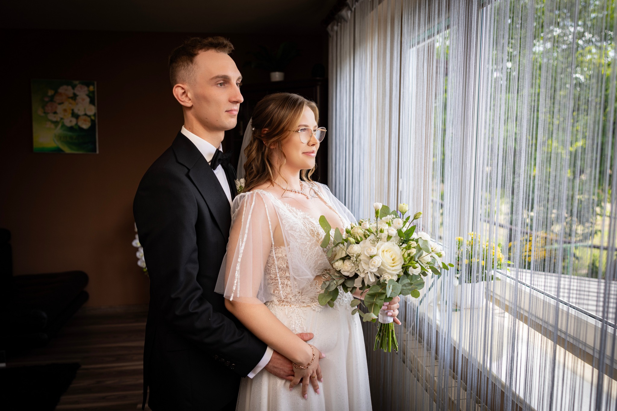Elegancka para młoda w Łodzi, on w smokingu, ona w sukni z bukietem, spoglądają przez okno z dekoracyjnymi zasłonami sznurkowymi, w tle zieleń drzew.