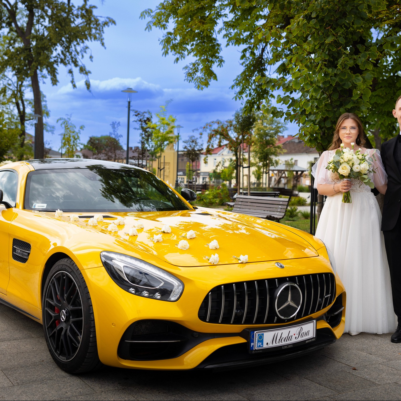 Żółty Mercedes z kwiatami, para młoda z bukietem na tle parku. Tablica rejestracyjna z napisem 'Młoda Para'.
