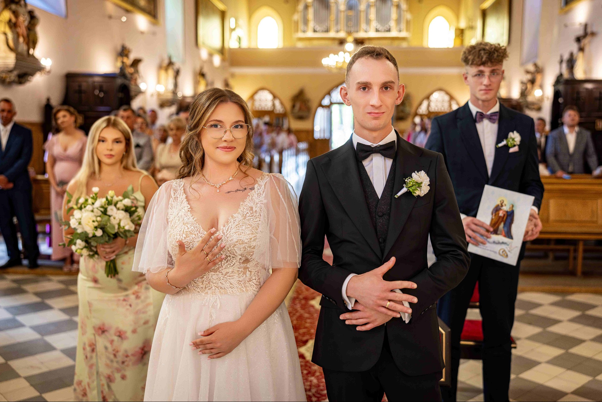 Para młoda w kościele, panna młoda w sukni z koronką, pan młody w czarnym smokingu z muszką. W tle goście i wnętrze kościoła, z dekoracjami i oświetleniem.