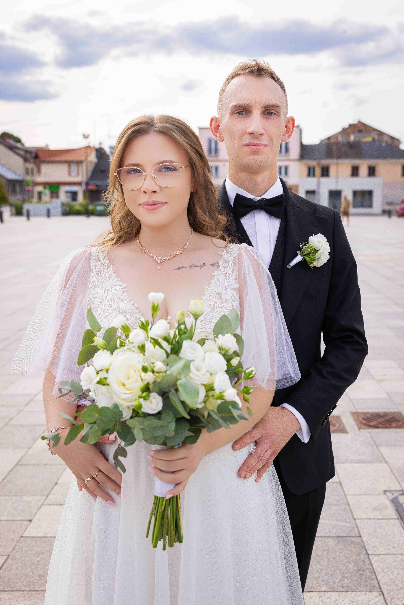 Elegancka para młoda w plenerze: panna młoda w okularach trzyma bukiet białych kwiatów, pan młody w czarnym smokingu. W tle budynki miejskie i pochmurne niebo.