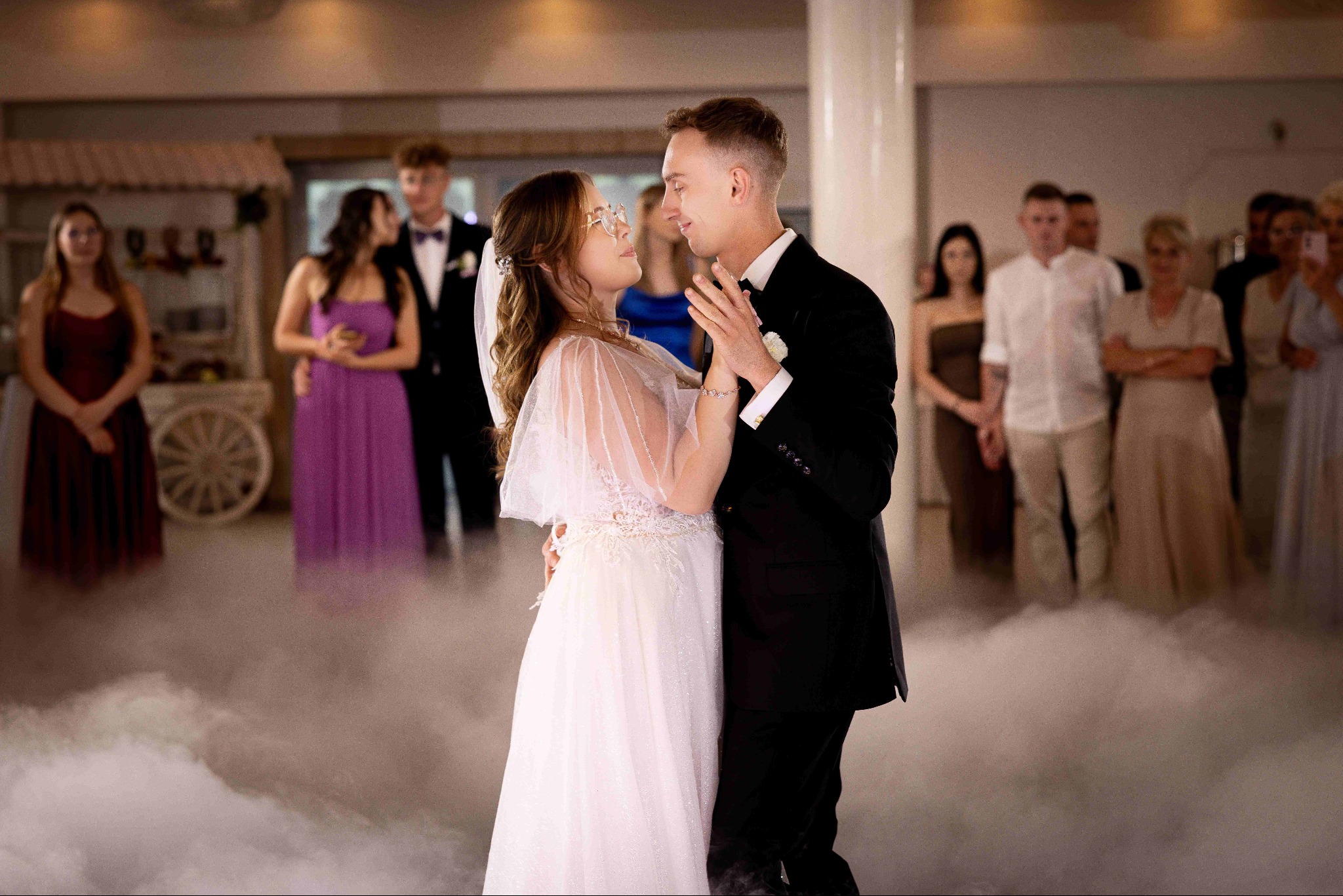 Para młoda w tańcu na tle gości weselnych i dekoracji. Panna młoda w okularach, dym na parkiecie, miękkie światło, uroczysta atmosfera.