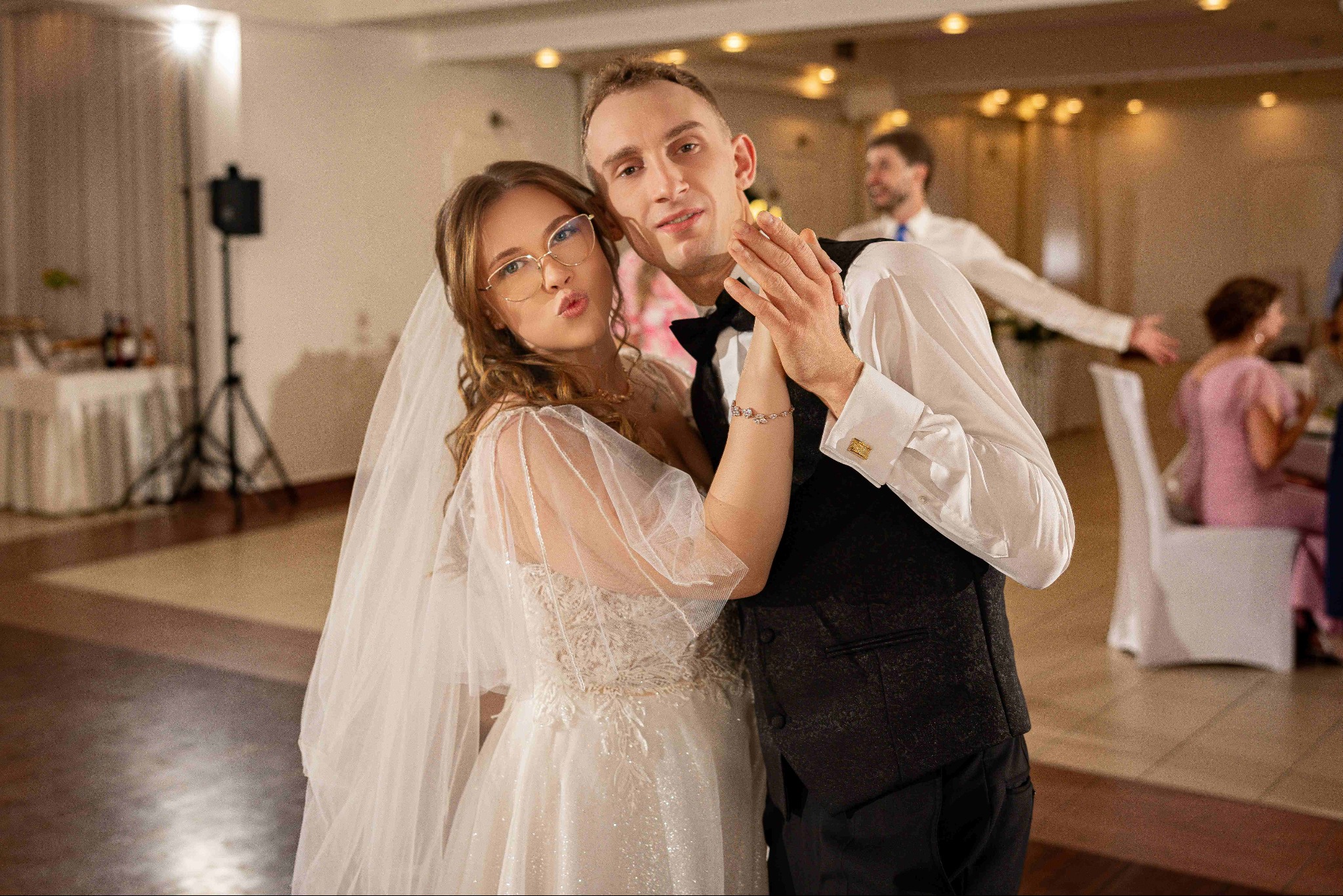 Para młoda w trakcie pierwszego tańca na sali weselnej. Panna młoda w okularach z zalotnym wyrazem twarzy. W tle goście i oświetlenie.