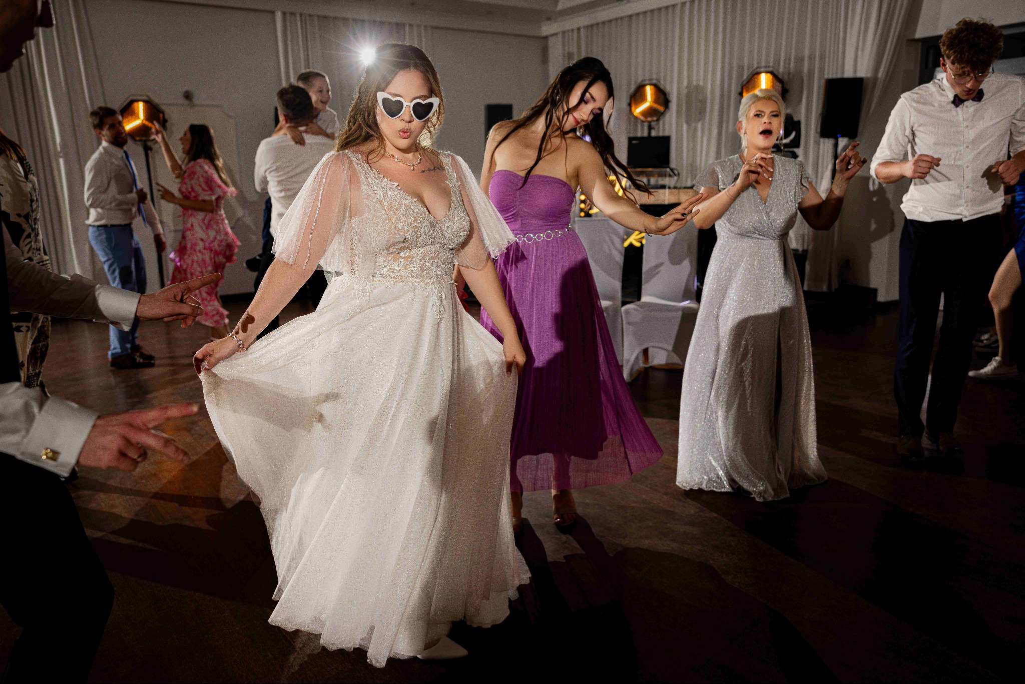 Roztańczona panna młoda w okularach w kształcie serc, w błyszczącej sukni, otoczona gośćmi weselnymi na parkiecie. W tle dekoracje świetlne i białe zasłony.
