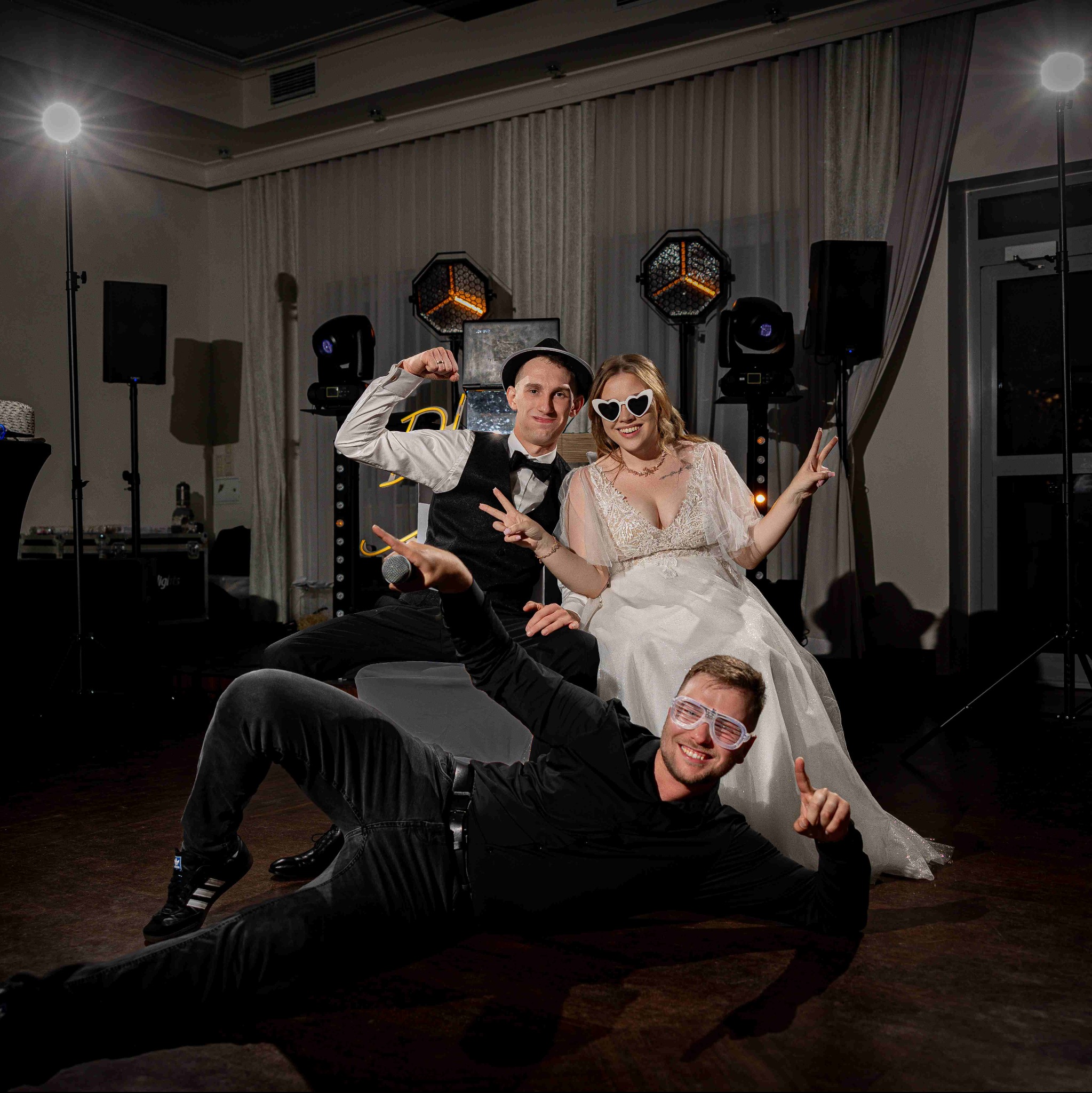 Radosna panna młoda w okularach w kształcie serc i pan młody w muszce pozują z fotografem na tle sprzętu oświetleniowego.