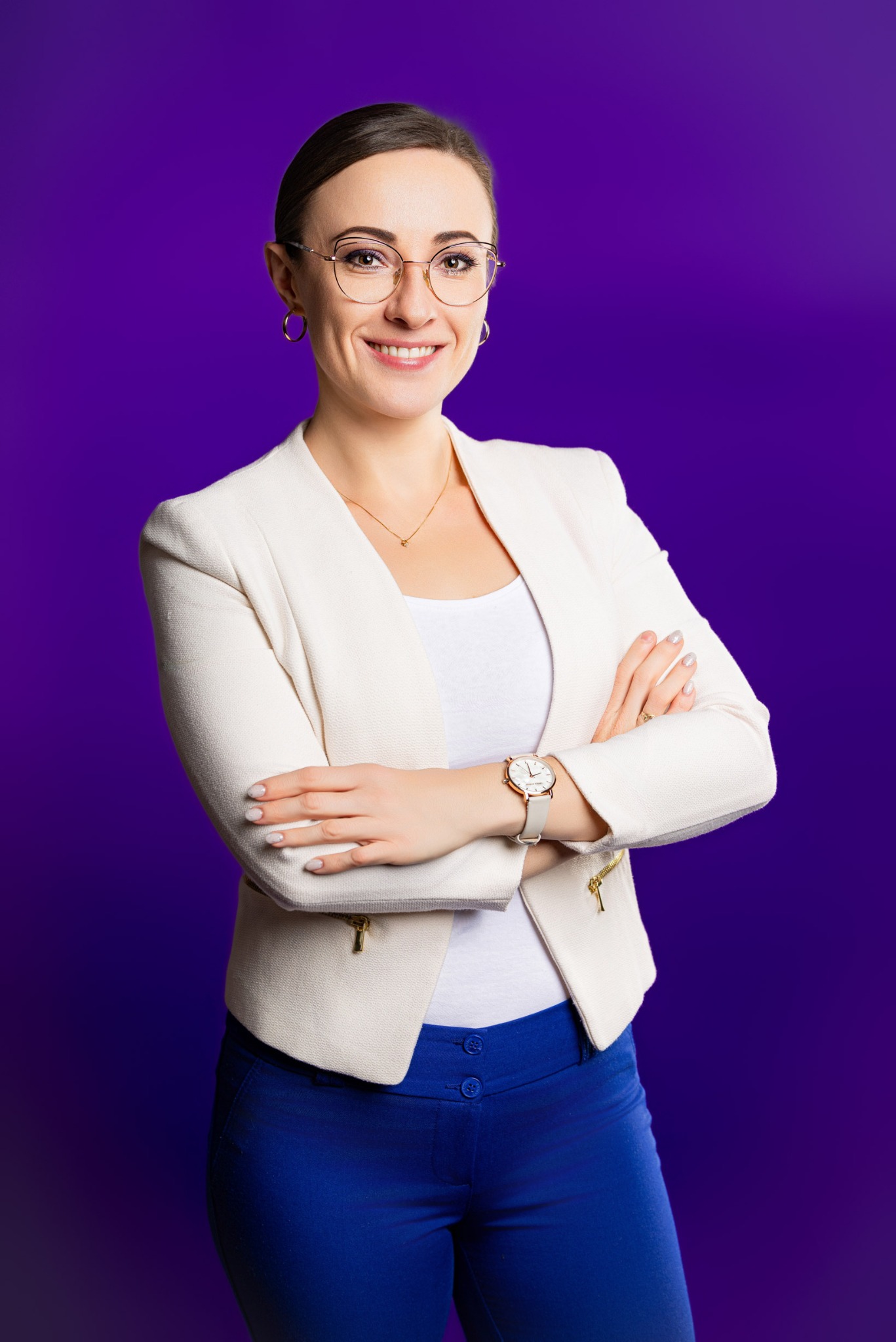 Portret uśmiechniętej kobiety w okularach, białym żakiecie i niebieskich spodniach na fioletowym tle. Skrzyżowane ręce z widocznym zegarkiem.