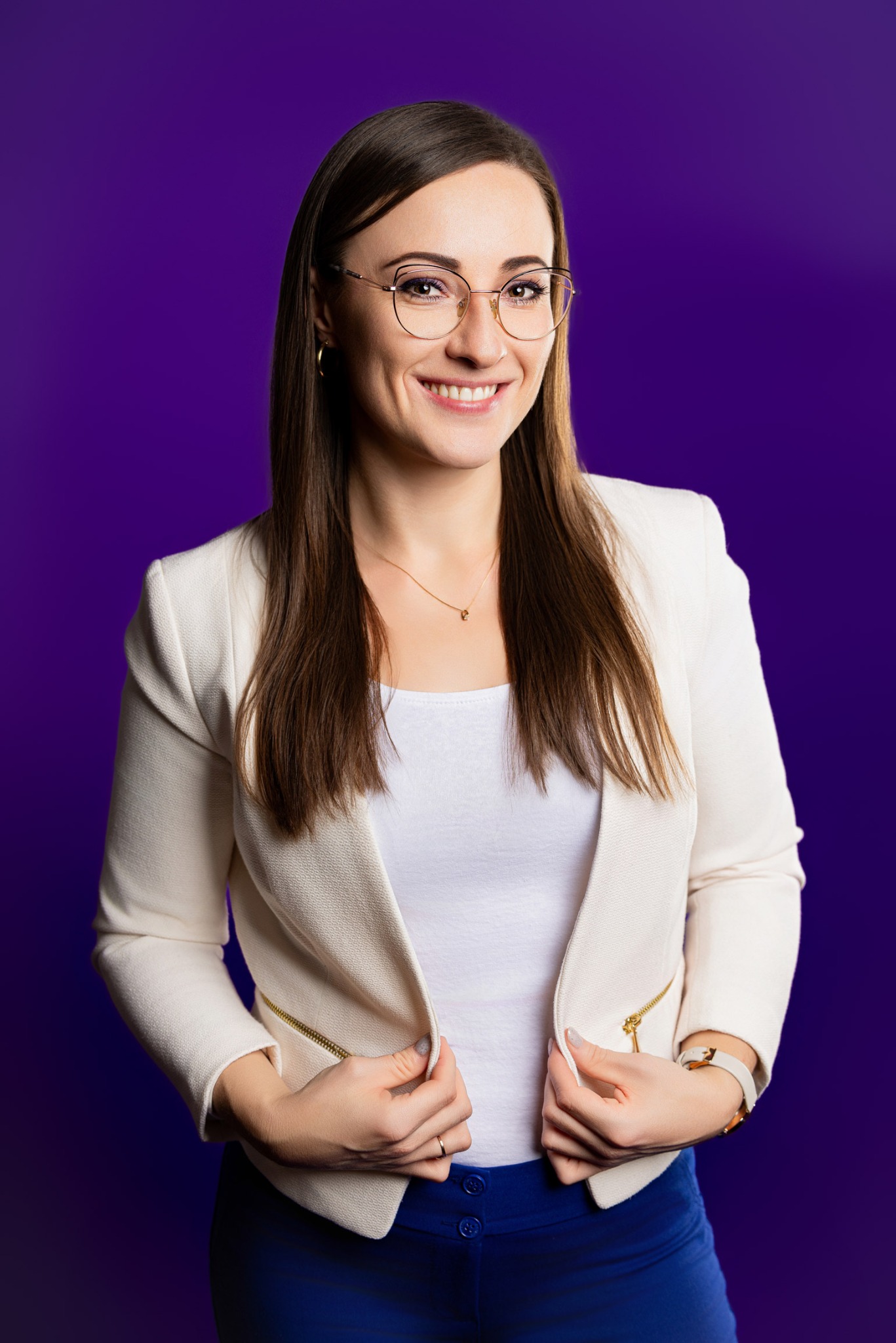 Portret uśmiechniętej kobiety w okularach i białym żakiecie na fioletowym tle, z delikatnym makijażem i subtelną biżuterią, prezentujący profesjonalny wizerunek.