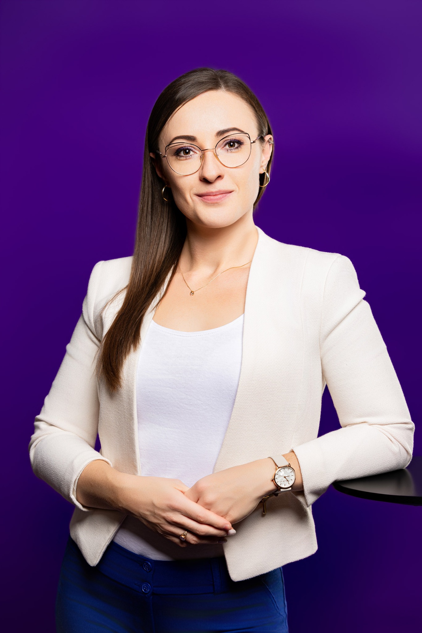 Portret kobiety w okularach, białej marynarce i niebieskich spodniach na fioletowym tle, opierającej się o czarny stolik. Subtelny uśmiech, profesjonalny wygląd.