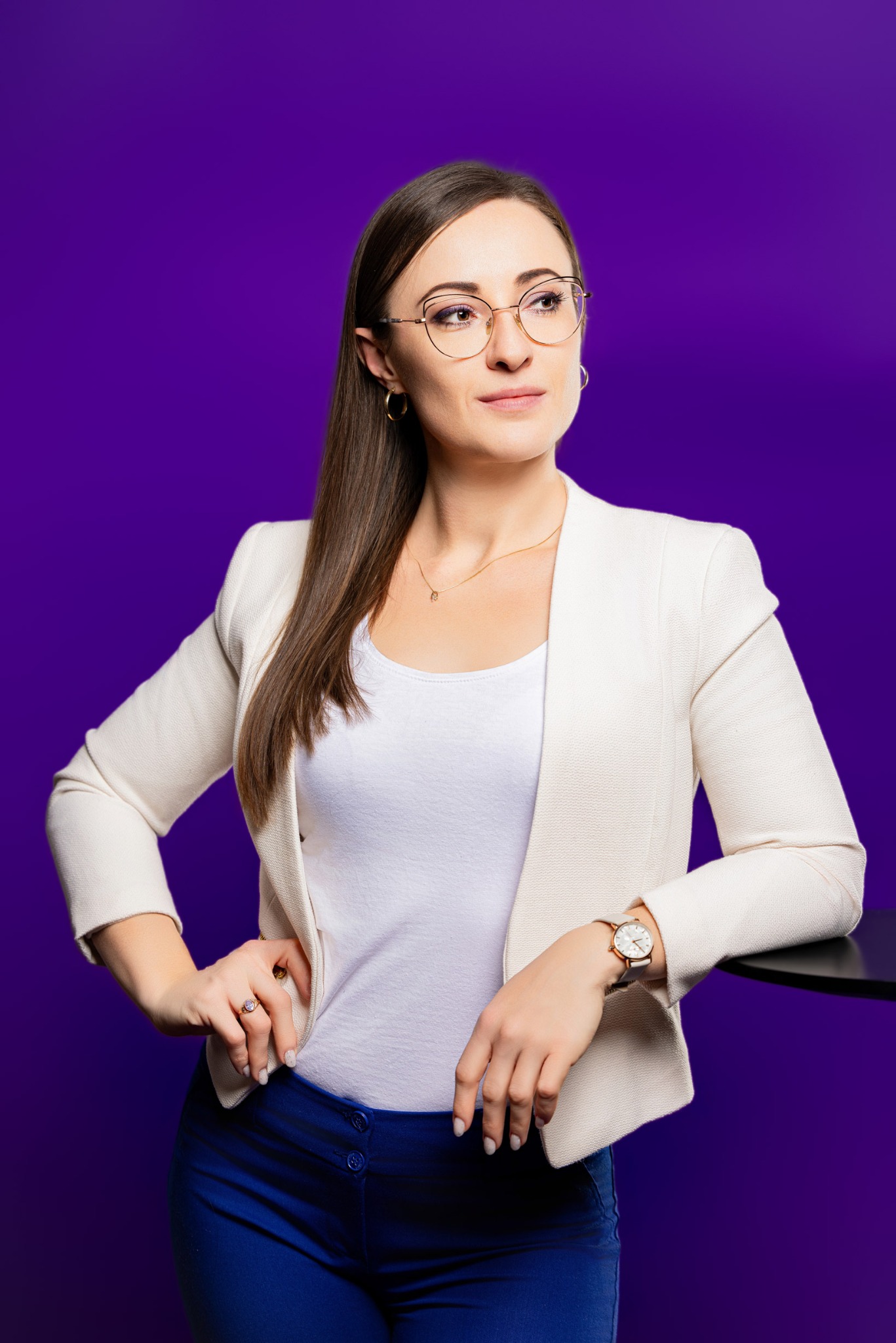 Portret kobiety w okularach, białej marynarce i niebieskich spodniach na fioletowym tle. Spogląda w bok, opierając się o czarny blat. Widoczny zegarek i pierścionek.