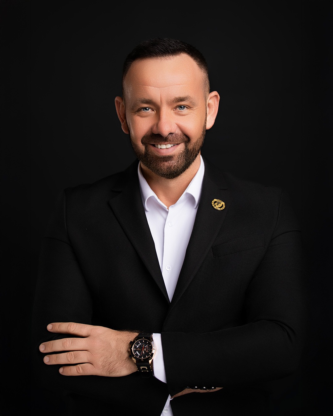 Portret uśmiechniętego mężczyzny w czarnym garniturze i białej koszuli na czarnym tle, z widocznym zegarkiem i przypinką w klapie.